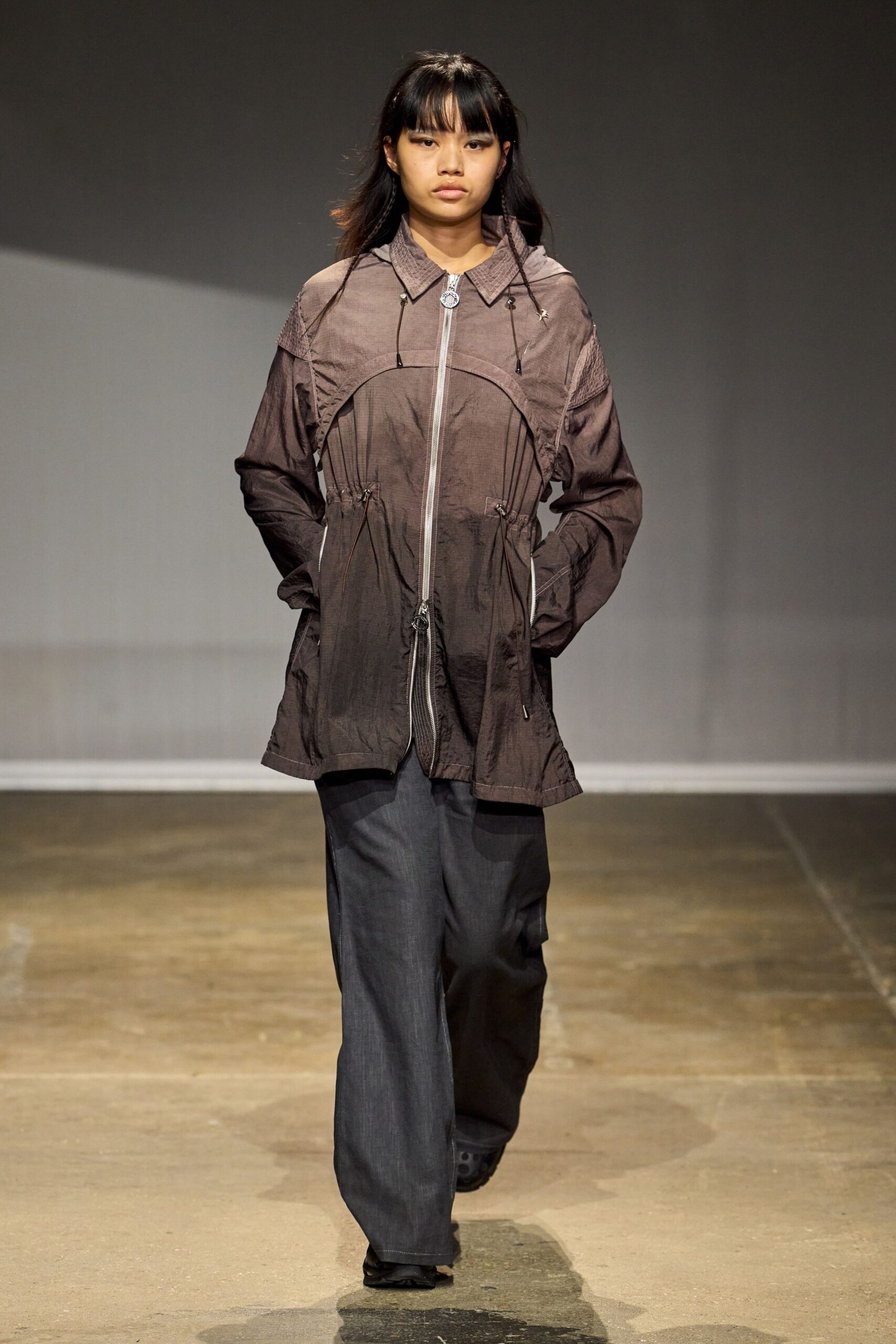 Smoky mauve crinkled shell jacket, curved seam across the chest and dangling toggles. Charcoal trousers with a dry denim look and soft break at the shoe. Black shoes peeking out blunt and rubbery. Walking across campus after rain, keys and receipts buried in every pocket.