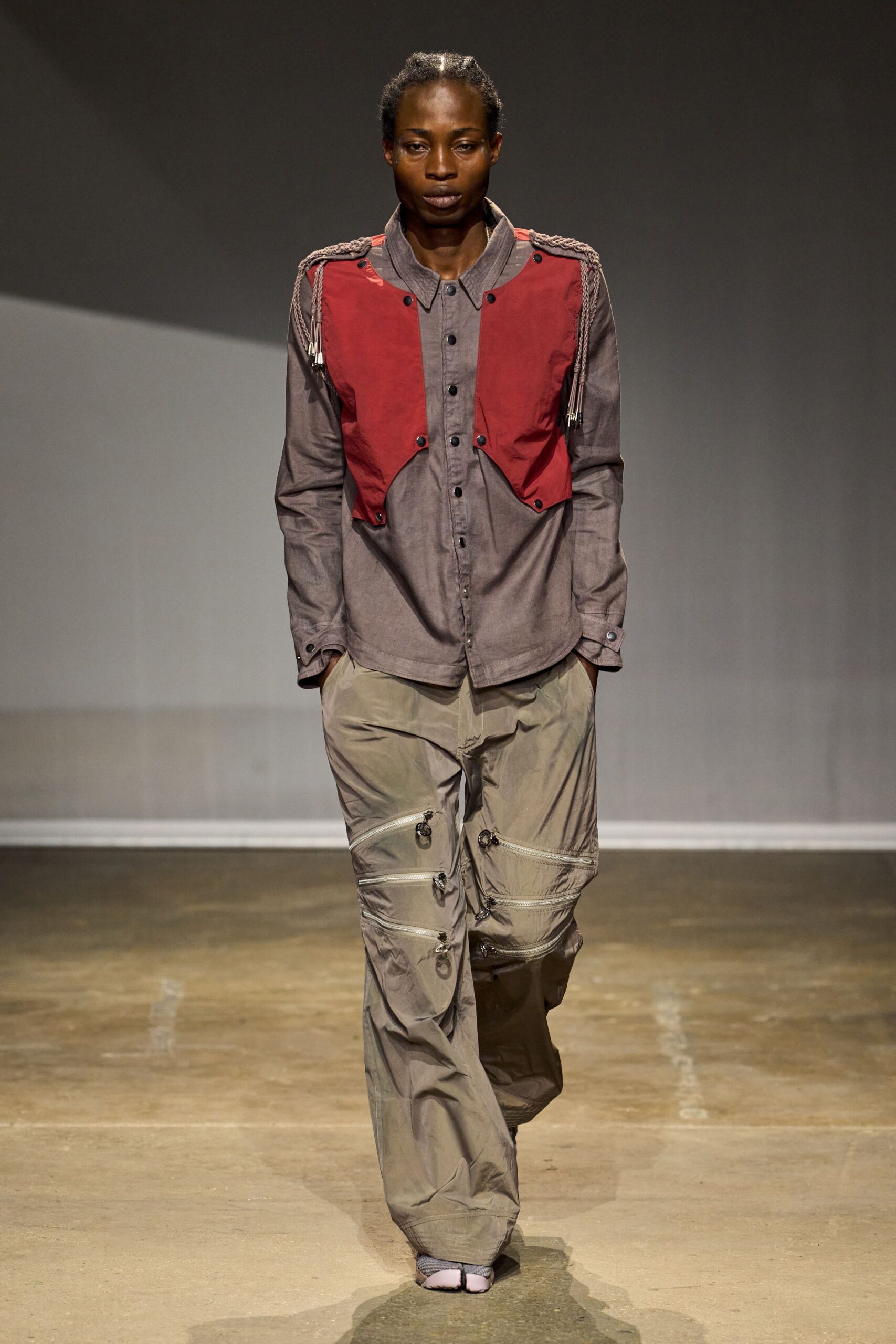 Brick nylon vest cut away at the waist, black snaps throughout. Taupe chambray shirt with a soft point collar and button cuffs. Dusty olive nylon, ring-pull zips banding both thighs. Split-toe slip-ons in pale mauve. Walking the long way across an airfield after the shuttle quit.