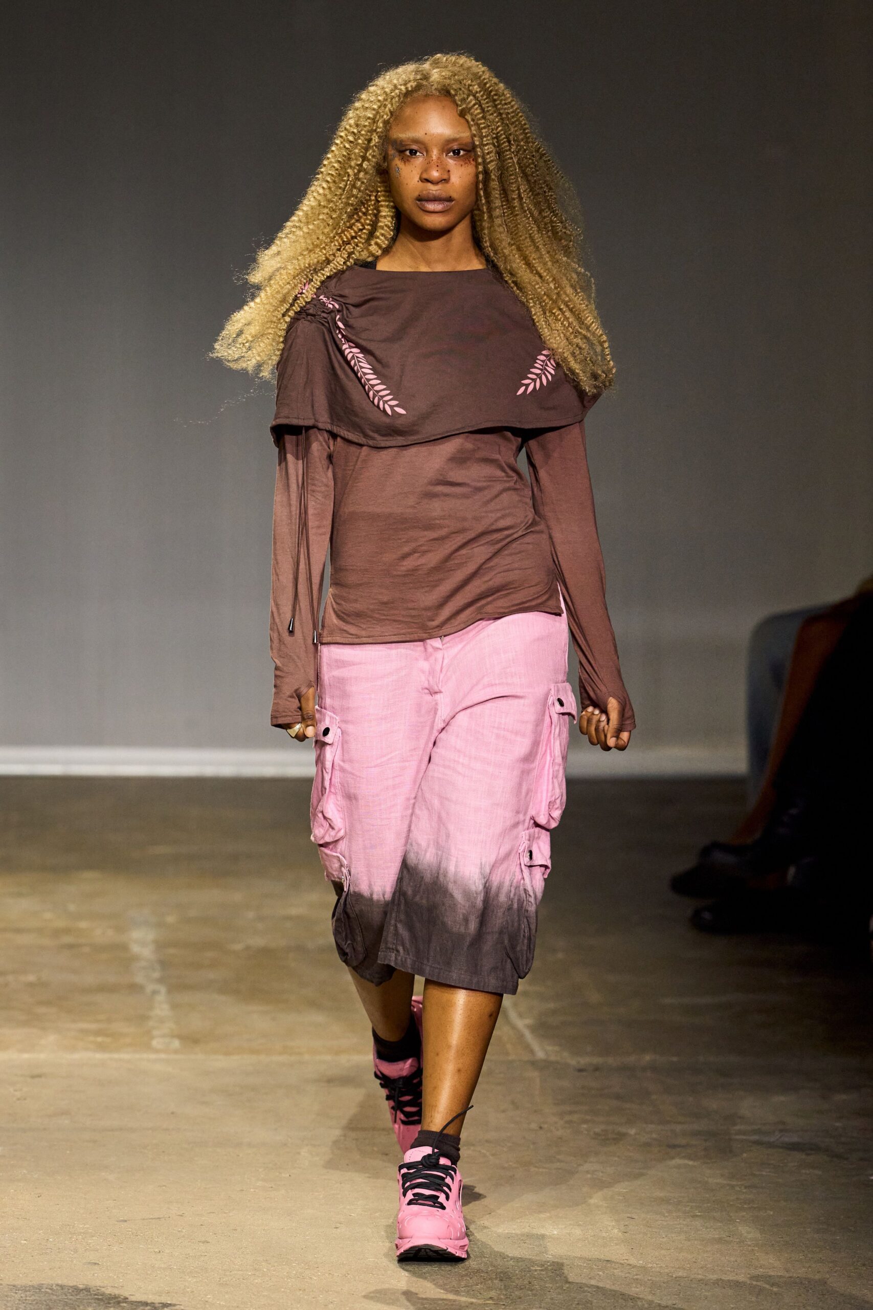 Chocolate jersey top with a cape-like yoke, ruched at one shoulder. Pink slub-weave bermudas dip-dyed to smoky brown, bellows pockets flaring. Bubblegum trainers with black laces and one loose ankle cord. Walking back from the corner shop as storm light turns everything mauve.