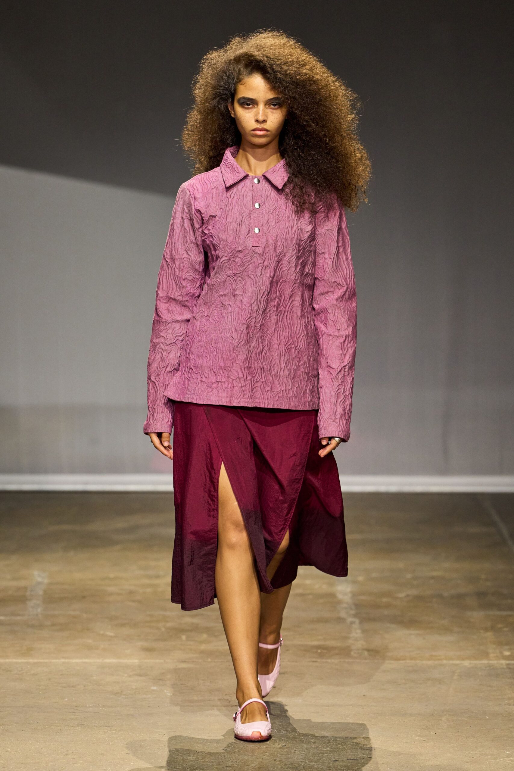 Crinkled mauve top with a pointed collar and three white snaps. Burgundy satin midi skirt, split high and wrapped off-center. Pale pink slingback flats with rounded toes. Walking across a hotel atrium before the flower delivery arrives.
