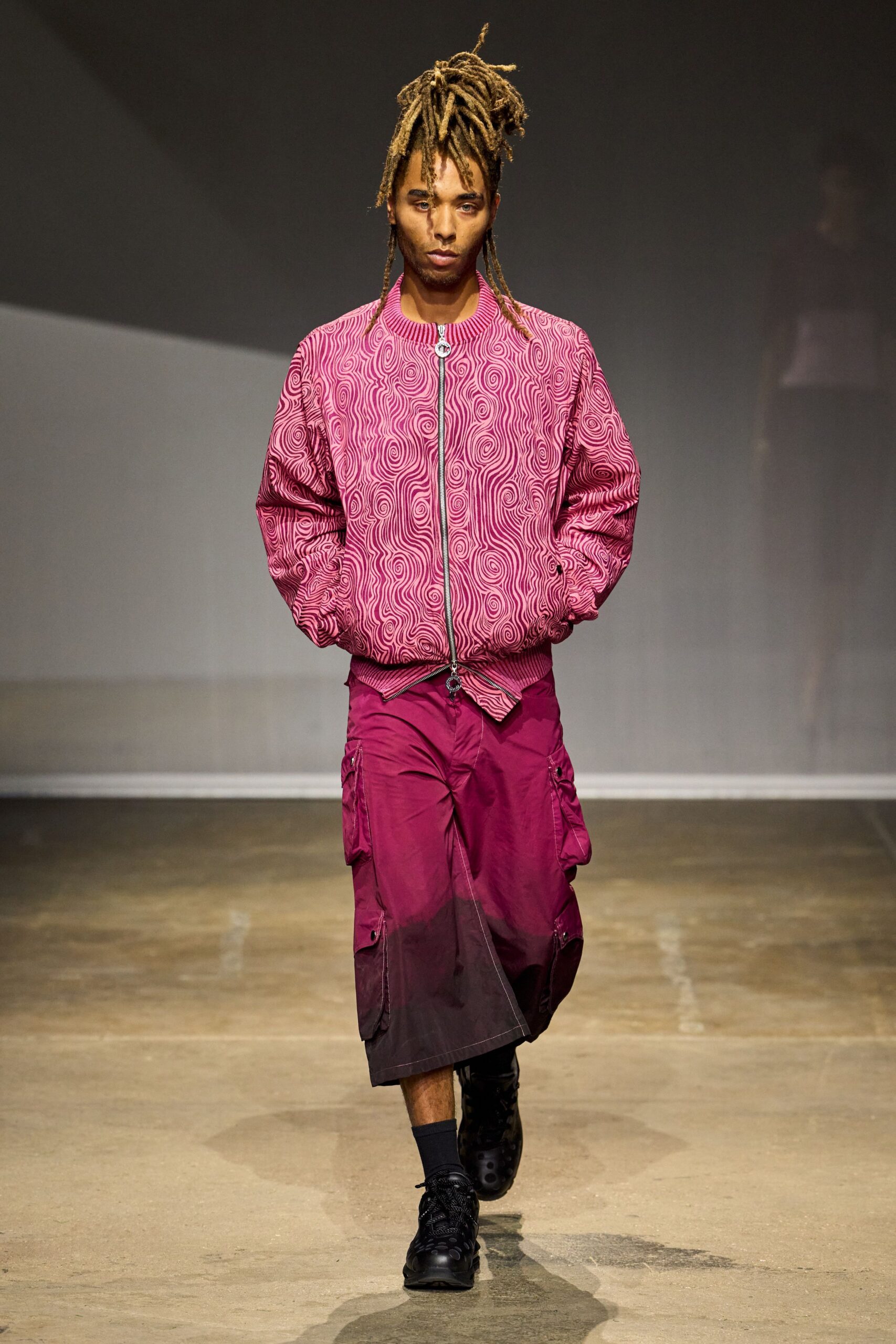 Pink jacquard bomber, maze-like swirls and a ring-pull zip. Magenta cargo shorts dip to plum at the hem, flap pockets low. Dark crew socks. Black trainers with punched-dot uppers. Walking back from the corner shop with melting ice pops in a heatwave.