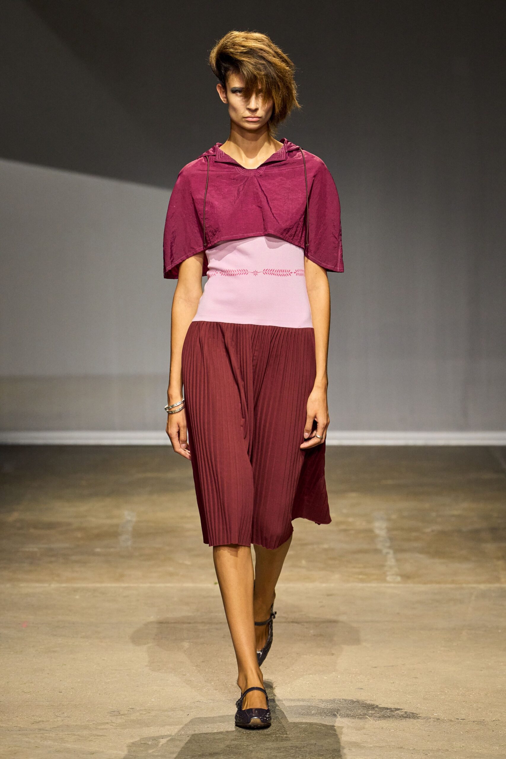Sheer mulberry capelet with a shallow hood and fine cord edging. Blush ribbed bodice dropping to a wine pleated skirt, tiny stitched motif at the waist. Stacked silver bangles. Dark plum mesh flats with a narrow strap. Walking out of a museum atrium after the rain has stopped.