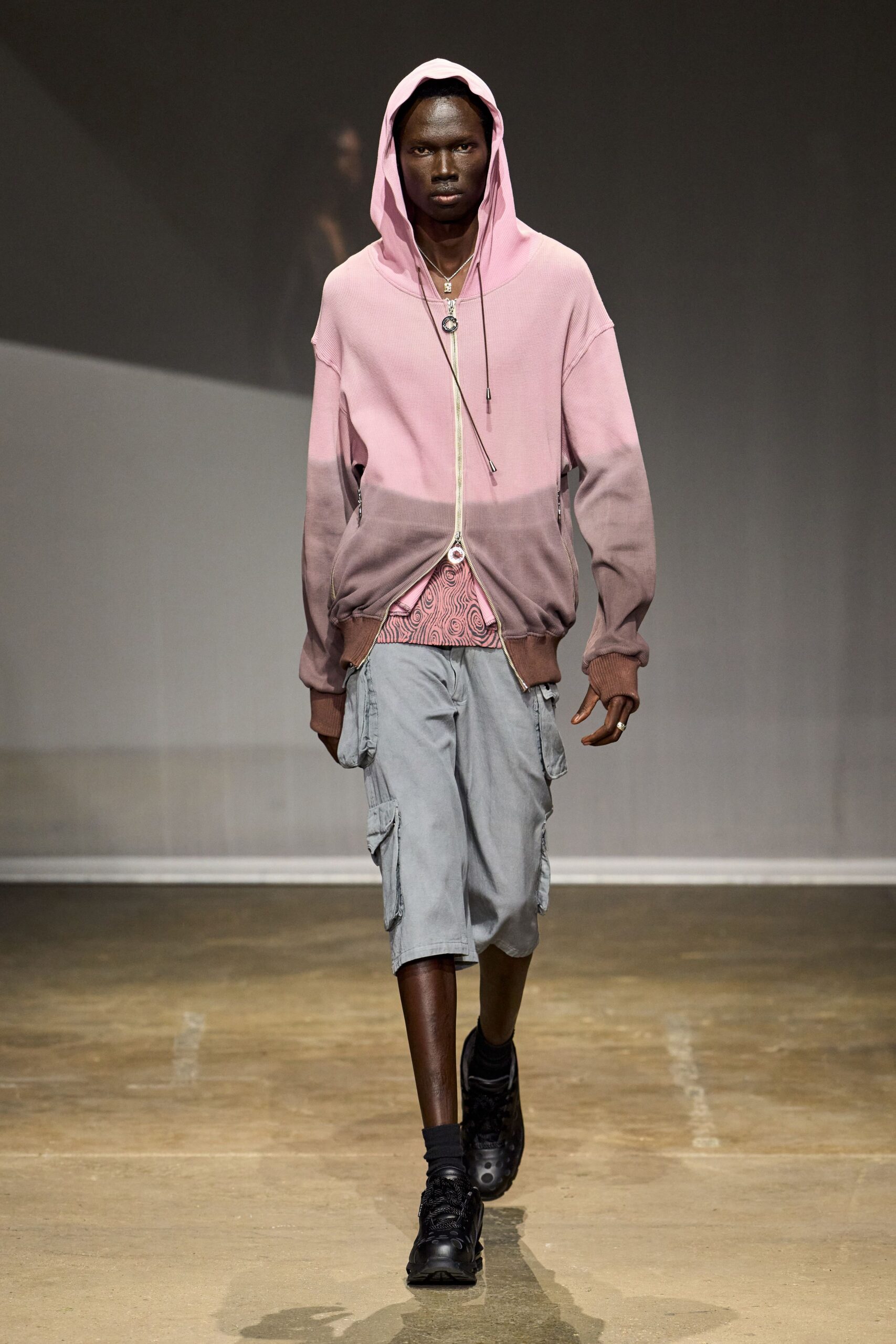 Pink waffle-knit zip hoodie fading to mauve, long drawcord tips hanging low. Swirl-print hem peeking through the opened zip. Washed pale-grey cargo shorts with flap pockets bumping at both thighs. Long cord pendant and tiny silver lock charm. Bulbous black sneakers. Walking to the corner shop just after rain, hood still up.