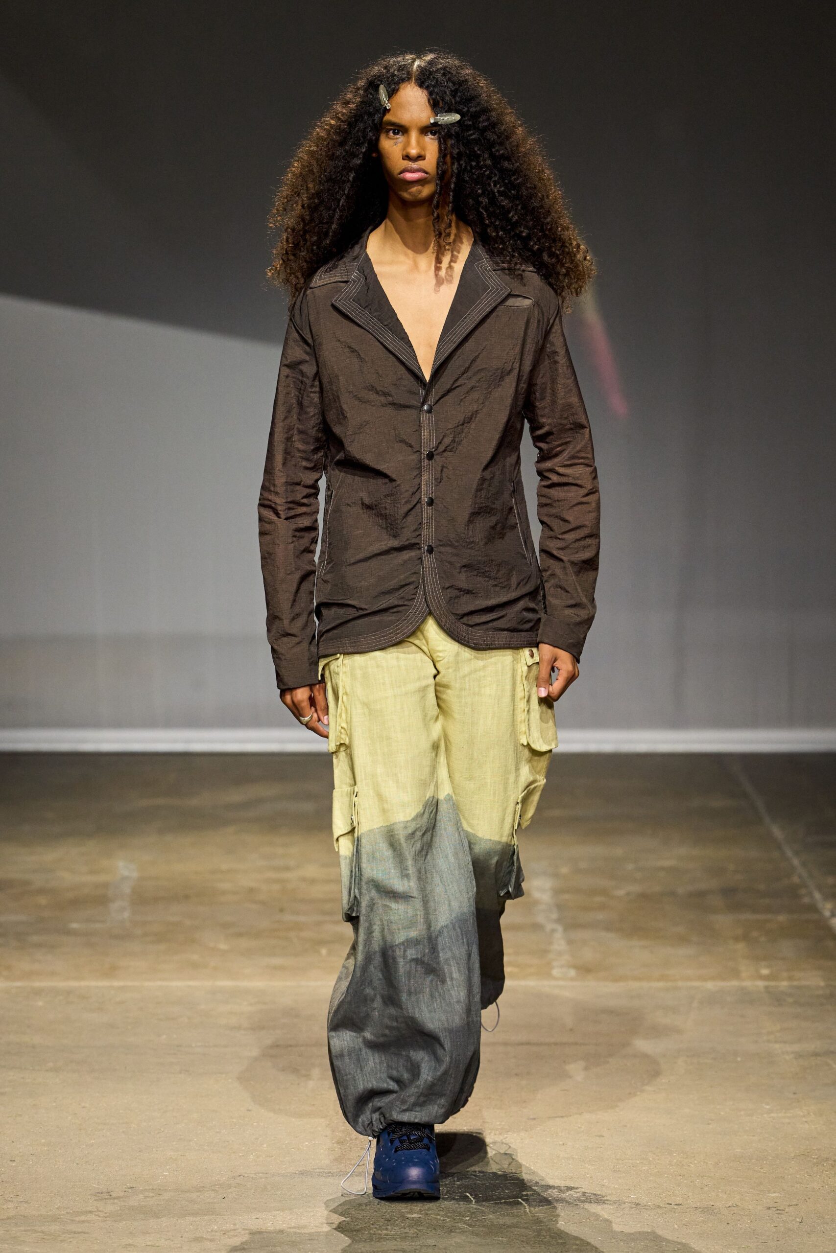Crinkled espresso blazer with pale topstitching tracing the notch lapels. Washed cargo pants fading from straw to charcoal, pockets flapping at the thigh. Cobalt trail sneakers with thick black laces. Walking across a fairground after rain, trouser hems already soaking through.