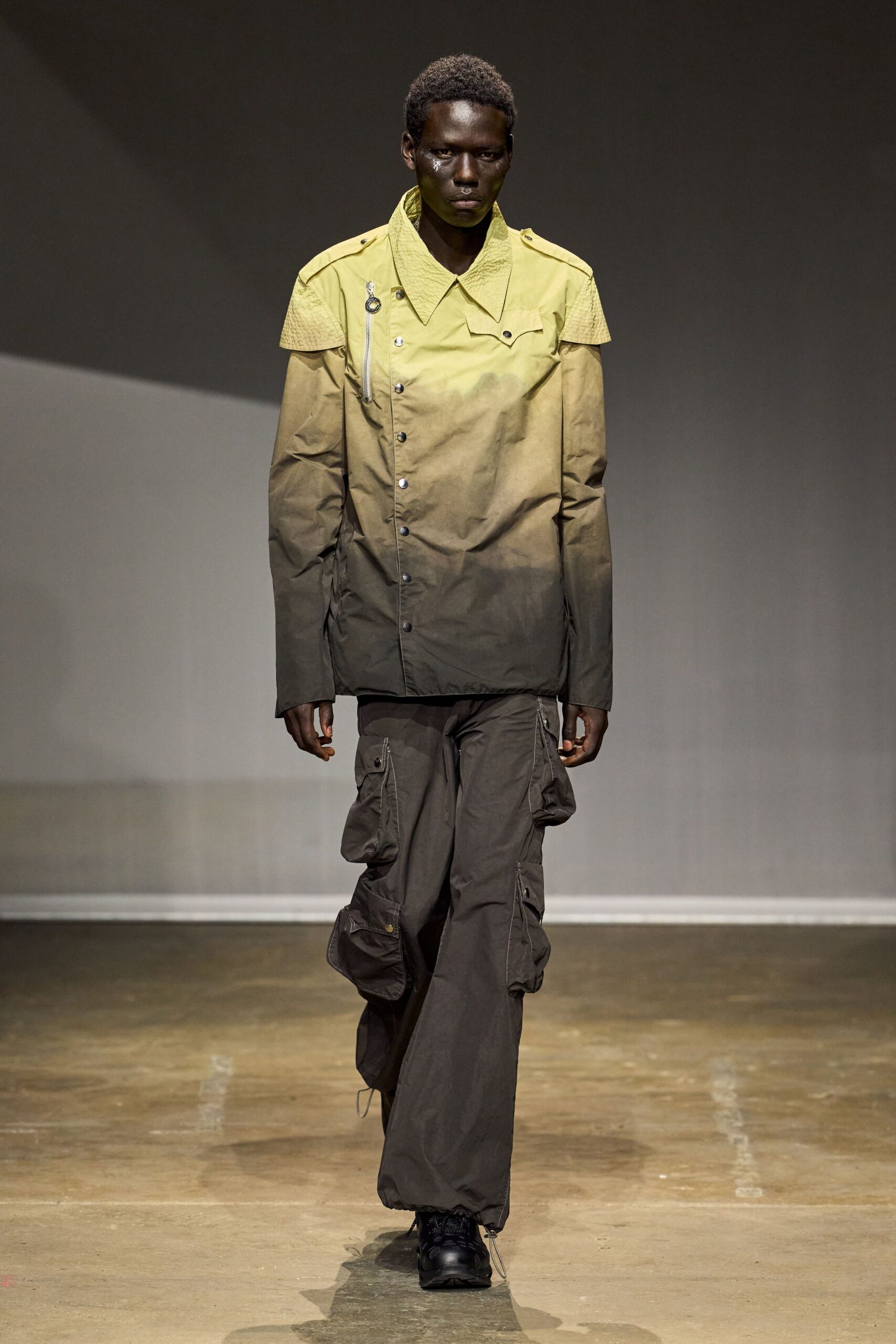 Sulphur-to-smoke snap-front field jacket, croc-embossed shoulder cape and ring zip pull. Charcoal nylon cargo pants with swollen flap pockets stacked down the legs. Black trail shoes, thick tread. Walking back from the reservoir with storm grit still in the air.