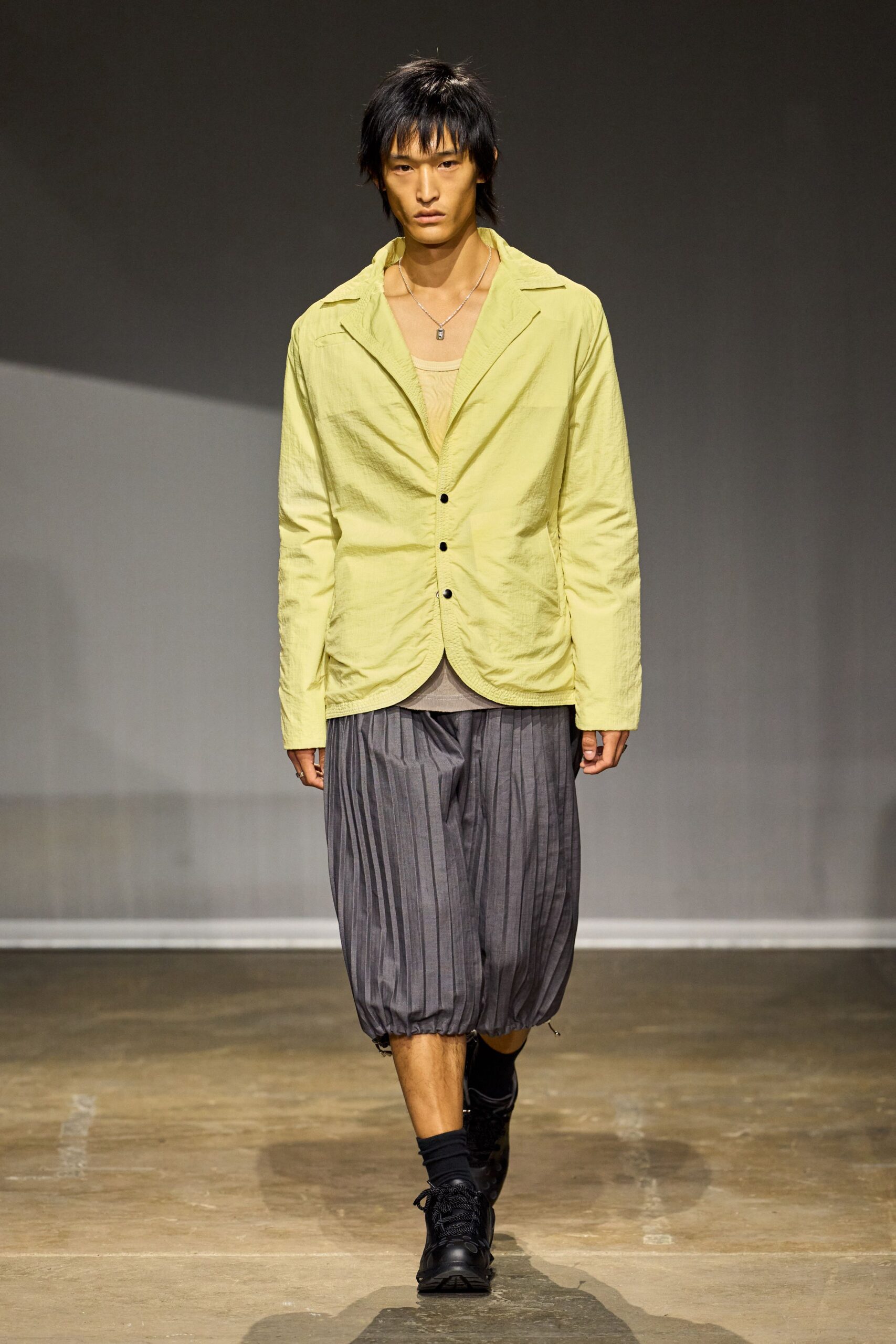 Crinkled citron jacket with black buttons and a softly collapsed notch lapel. Sheer beige tank peeking under the open front. Charcoal pleated balloon shorts, cinched tight at the hem. Small silver pendant on a fine chain. Black low-top sneakers with thick rope laces. Walking to buy cold fruit just before the heat fully sets in.