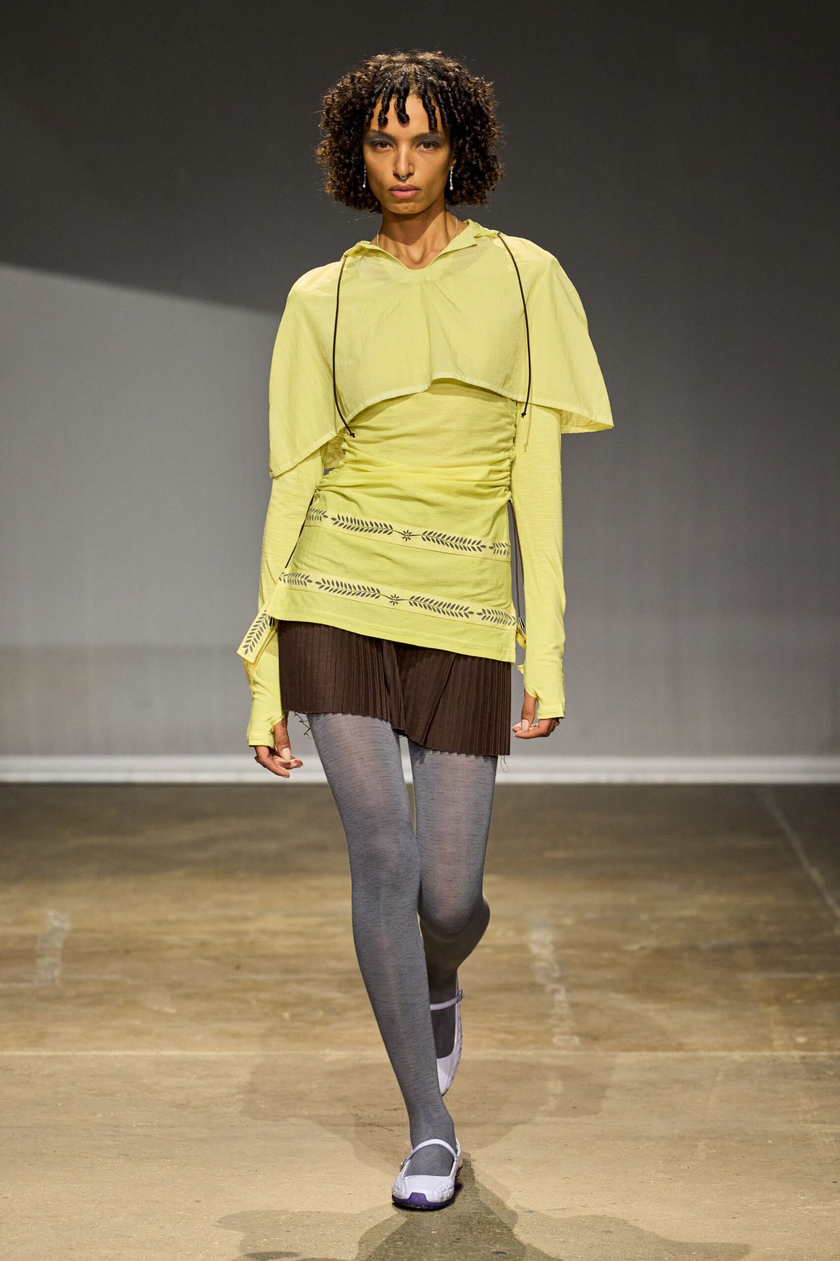 Pale citron voile cape layer with black drawcords hanging loose. Ruched citron jersey tunic, thumbhole sleeves and tiny cutout borders. Chocolate micro-pleats peeking unevenly below the hem. Flat grey tights. Lilac sport soles and square toes. Walking to the corner shop after rain, still in yesterday's eyeliner.