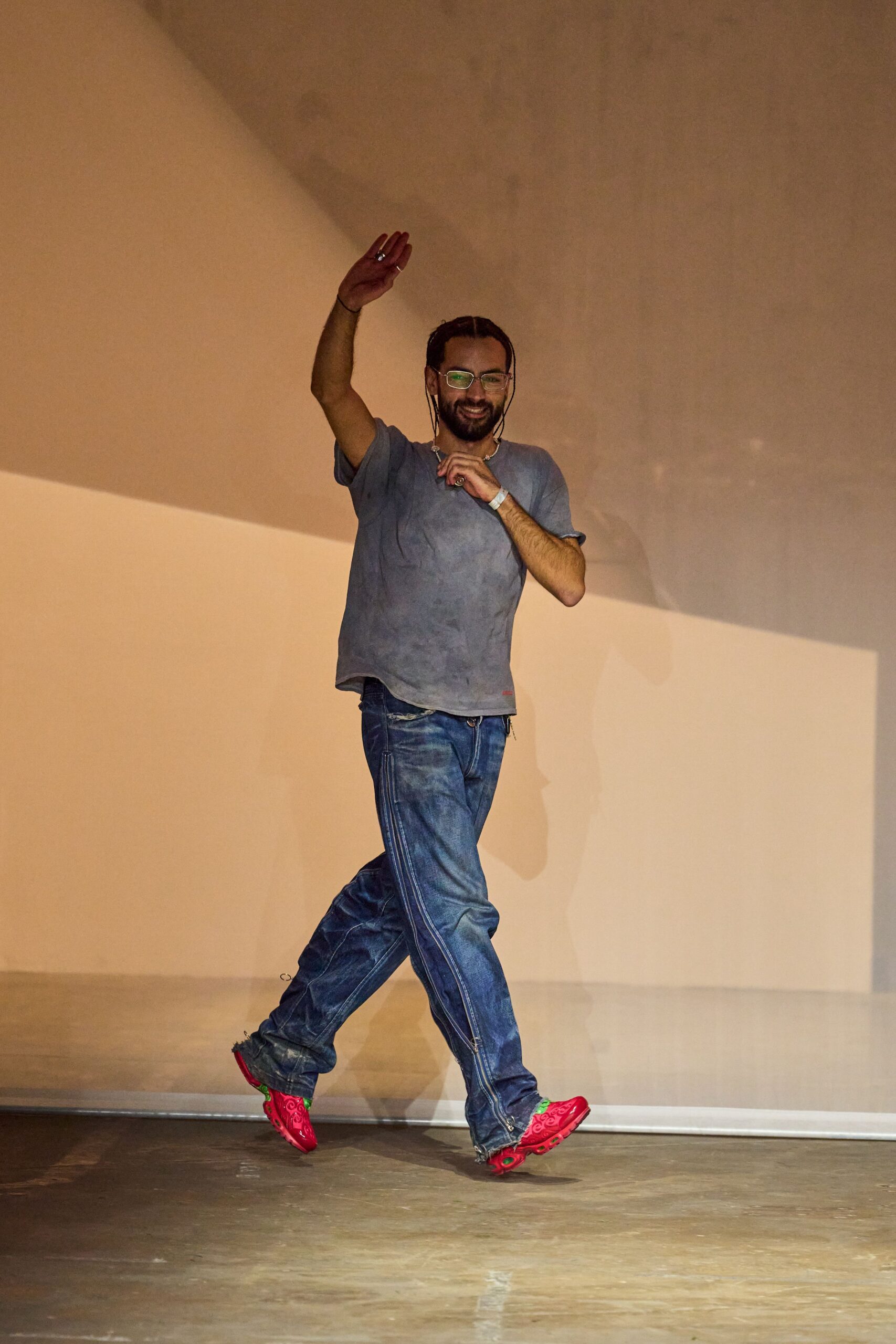 Clear frames with a black cord dropping past the jaw. Washed slate tee with rolled sleeves and a slightly curled hem. Loose blue jeans with double side seams and frayed zip hems. Hot-pink trainers with green lining and paisley-patterned uppers. Waving from the DJ booth once the lights finally come up.