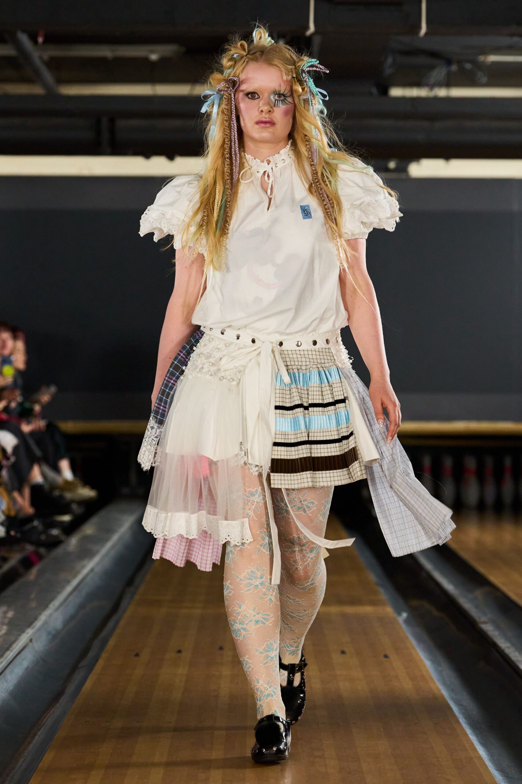 Ivory blouse with puff sleeves and broderie anglaise cuffs. Patchwork wrap skirt mixing tulle, plaid shirting and pleated striped panels. Sheer tights stitched with pale blue floral lace. Black patent Mary Janes with chunky silver buckles. Walking from a church fete into the school hall before the bunting comes down.