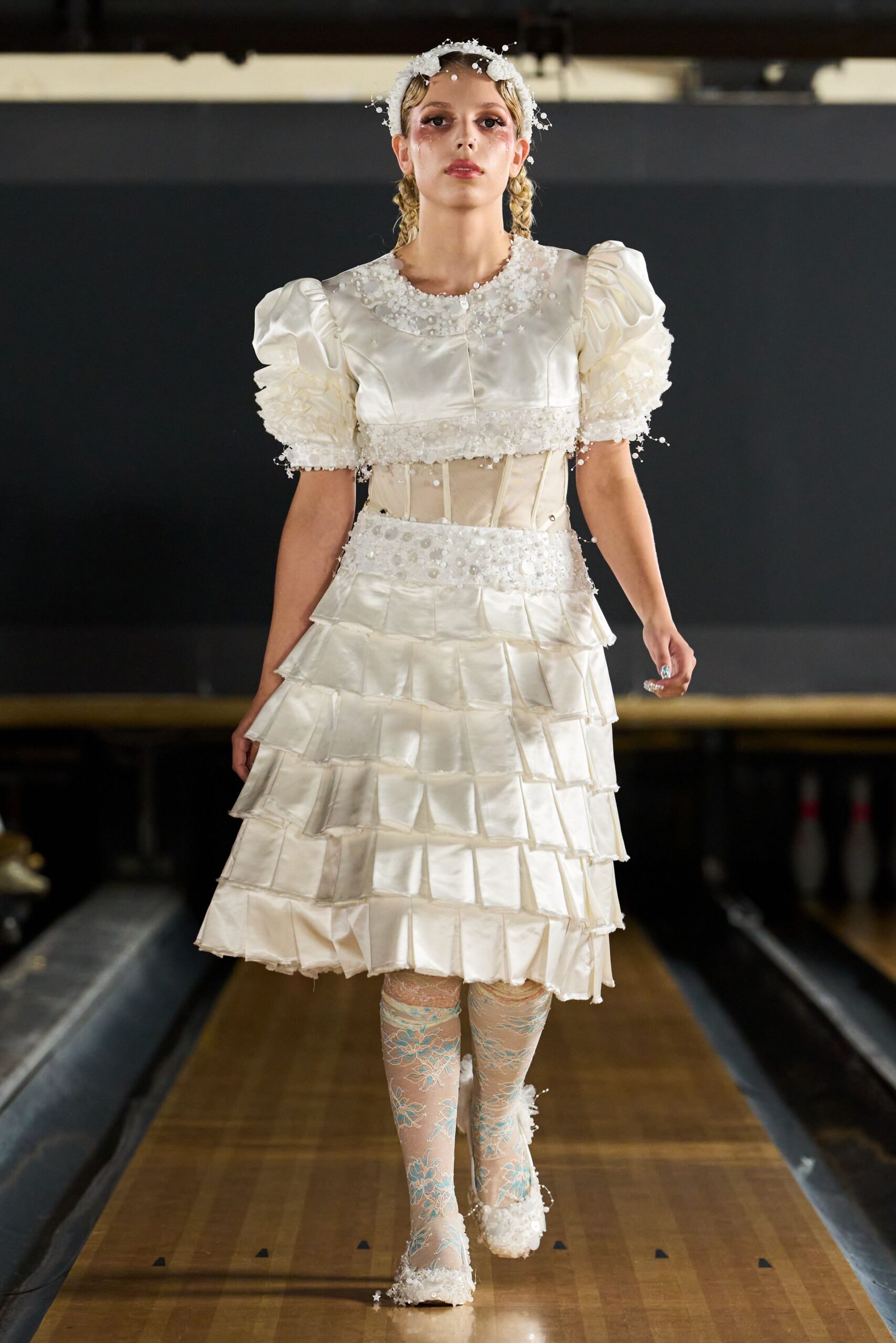 White floral headband dotted with wiry pearl sprays. Ivory satin dress with puff sleeves, boned mesh midriff and tiered square flounces. Nude lace tights washed with pale blue floral tracery. Square-toe white shoes crusted at the edges with pearly beading. Walking through a church hall after the cake has already been cut.