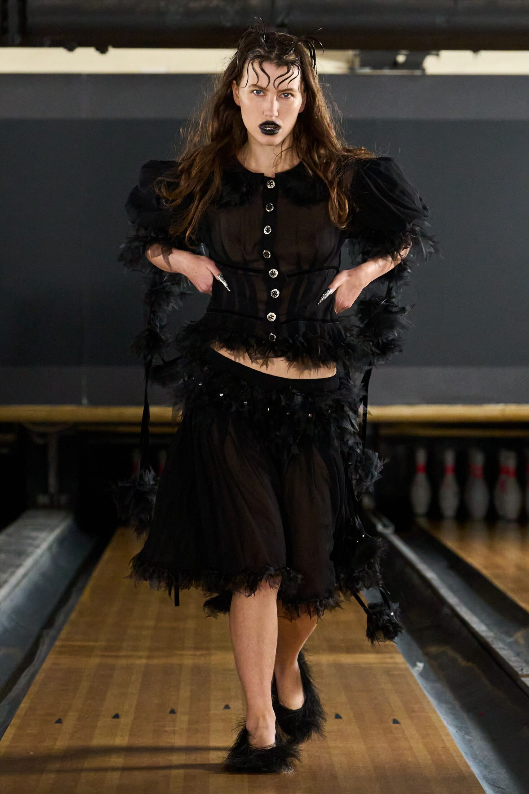 Sheer black chiffon blouse with puff sleeves and marabou-trimmed edges. Rhinestone buttons running straight down the transparent front. Sheer black skirt with a low waist and feathered hem. Black pumps tufted with fluffy marabou at the toe. Bowling alley hush, one lane still glowing after midnight.