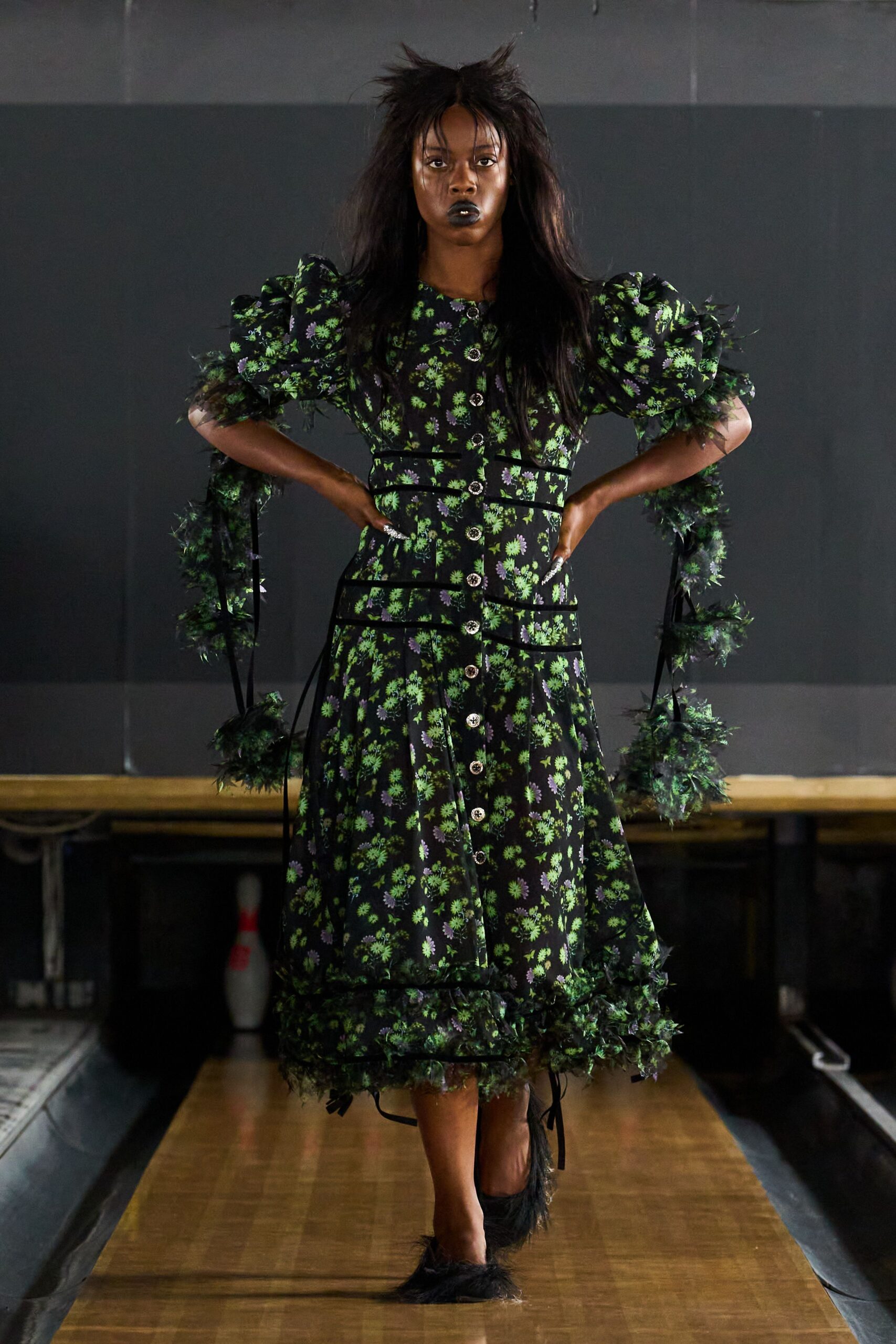 Black floral dress with puff sleeves, rhinestone buttons, and feathery trim trailing from the arms. Black flats tufted with shaggy marabou at the toes. School auditorium after hours, one bowling pin still standing by the wall.