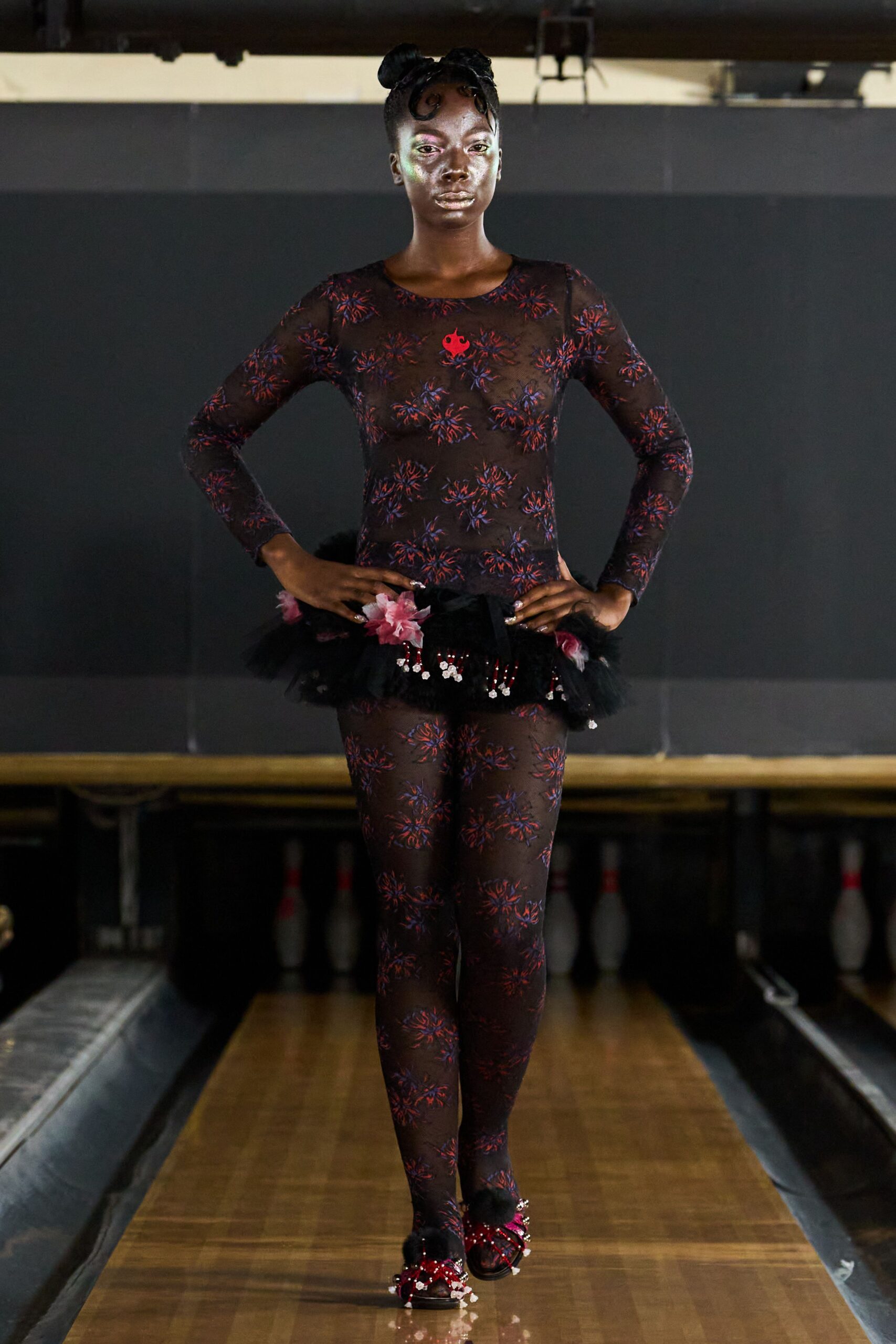Sheer black mesh bodysuit scattered with red and violet floral bursts. Black tulle peplum mini, pinned with pink blossoms and dangling beads. Black sandals tufted with pom-poms and crimson crystal fringe. At the bowling alley after midnight, still dressed for the recital.