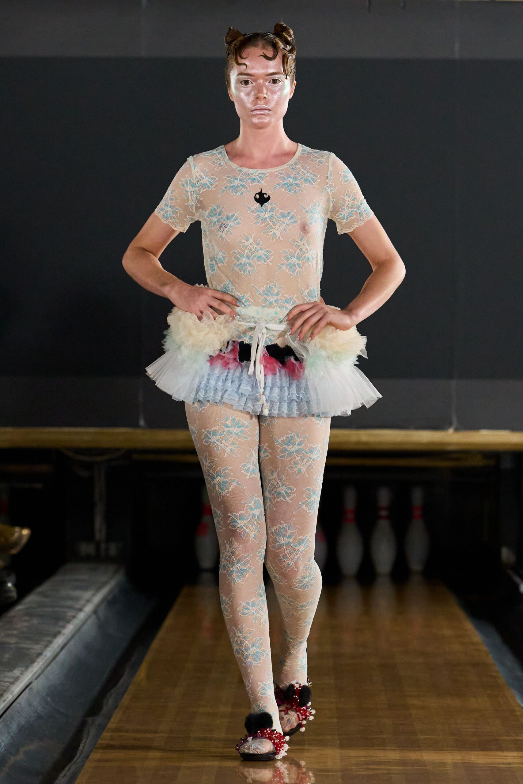 Sheer nude mesh tee embroidered with powder-blue floral lace. Tiered tulle peplum with ruffled blue lace and pink tufts. Matching floral lace tights, the motif repeating uninterrupted to the feet. Beaded red flats with black pompom toes. Between bowling lanes after midnight, still dressed for a toy-box recital.
