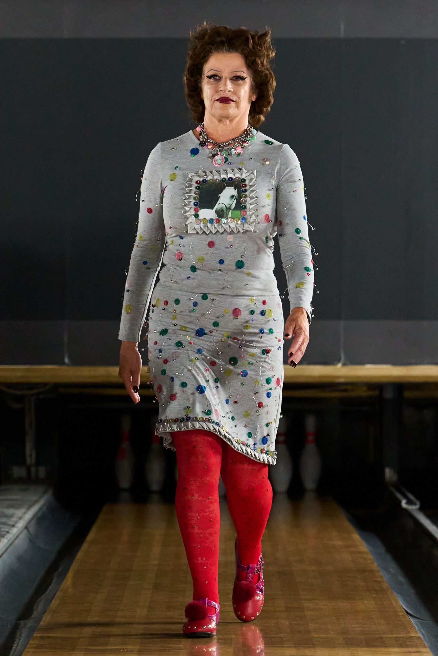 Pale grey jersey dress speckled with sequins, horse picture framed in folded spikes. Chunky charm necklace with a pink cameo hanging at the throat. Tomato red tights dotted with tiny rhinestones. Glossy red Mary Janes with pompom straps and metal studs. At the small-town bowling alley before the birthday sheet cake comes out.