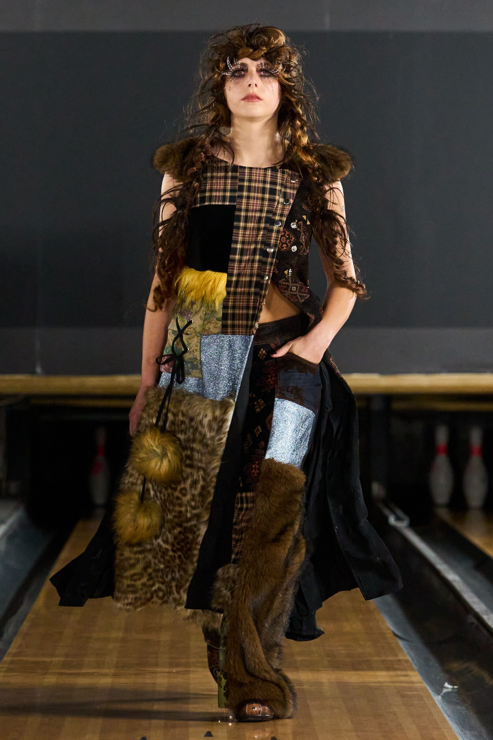 Spidery crystal fringe fanning from the eyes into the hairline. Asymmetric patchwork dress in tartan, glitter knit, brocade and fur panels. Mismatched patchwork boots with shaggy brown fur climbing past the knee. Walking the bowling alley after midnight, lanes still warm underfoot.