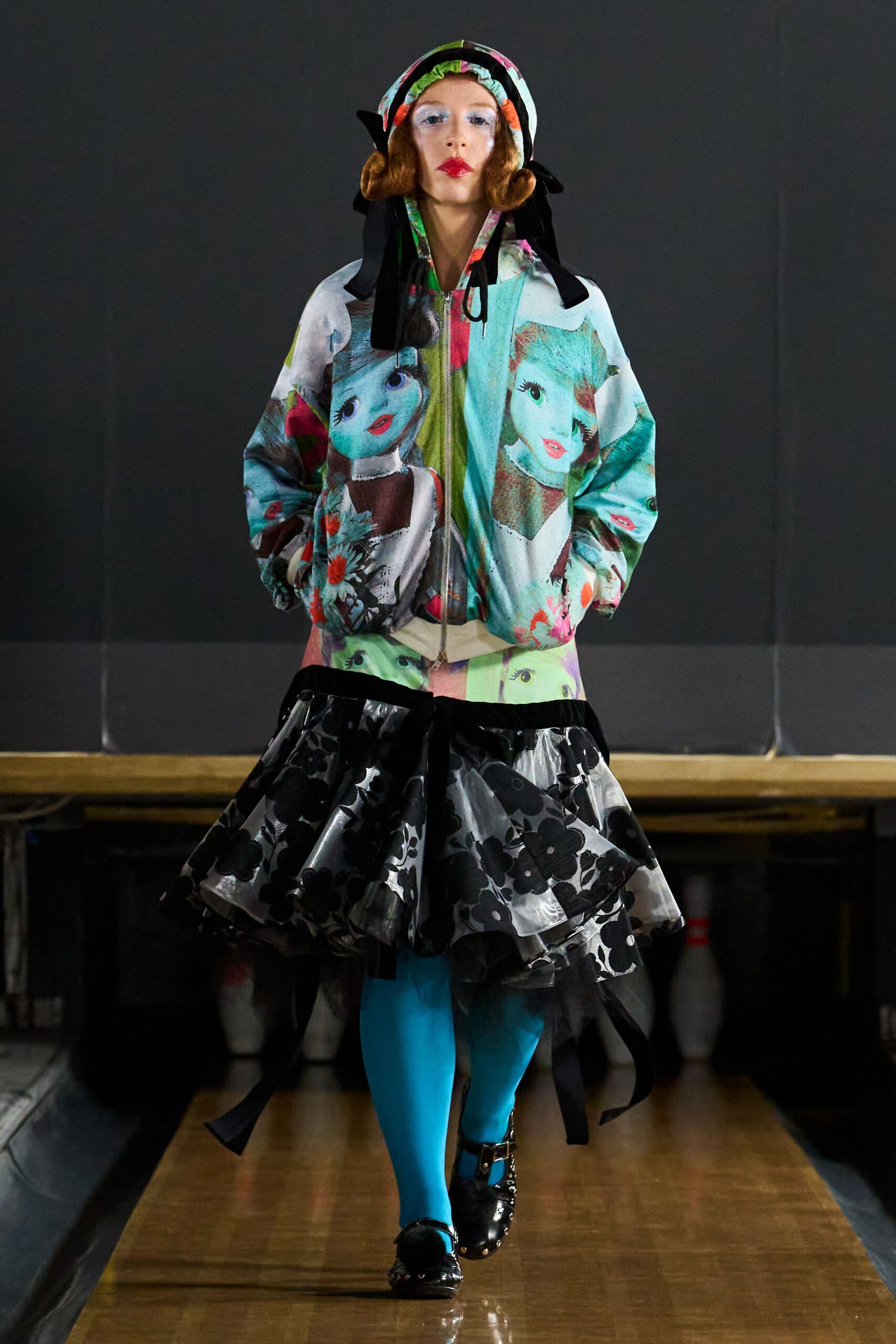 Printed bonnet with ruched edge and long black ribbon ties. Zip-front hooded jacket printed with washed-out doll faces. Tiered clear vinyl ruffle skirt flocked with black flowers. Opaque turquoise tights. Black patent Mary Janes with a furry pom at one toe. School auditorium after hours, bowling pins stacked under the stage.