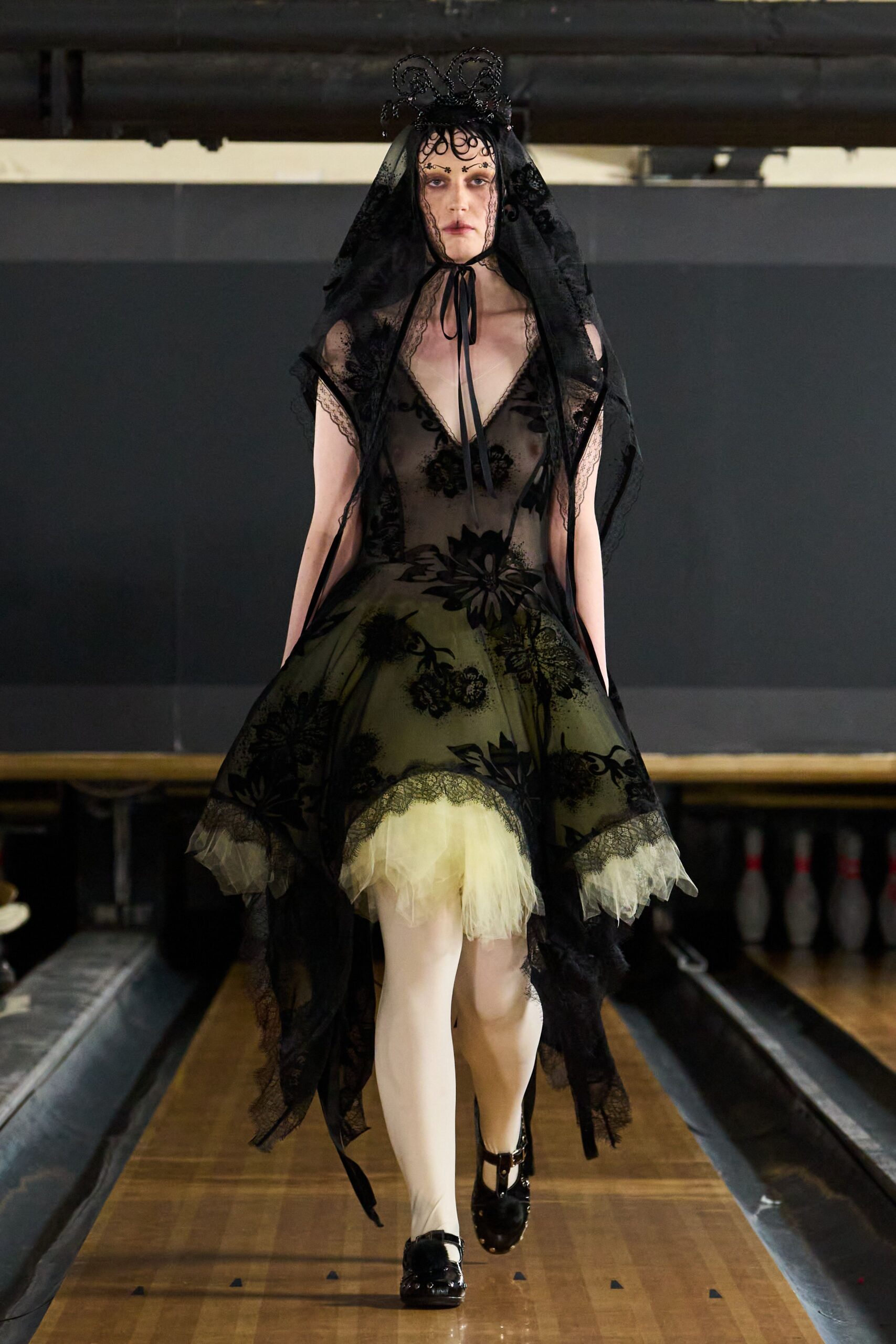 Black looped wire crown under a long lace-edged mourning veil. Sheer black floral flocked dress, handkerchief hem lifted over butter tulle. Opaque ivory tights. Patent black Mary Janes with a blunt strap across the instep. Crossing a bowling alley after the wake, lanes still glowing.