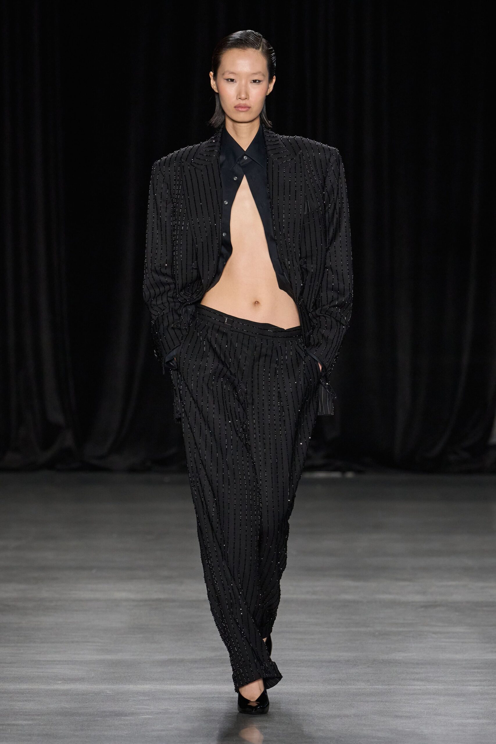 Black blazer with padded shoulders and glittering bead pinstripes. Black poplin shirt buttoned only at the collar, cut to a deep front void. Low-slung matching trousers, pleats collapsing into a twisted wrap front. Black patent pumps, sharply pointed. Whatever you wear home when dawn catches you leaving the hotel bar.