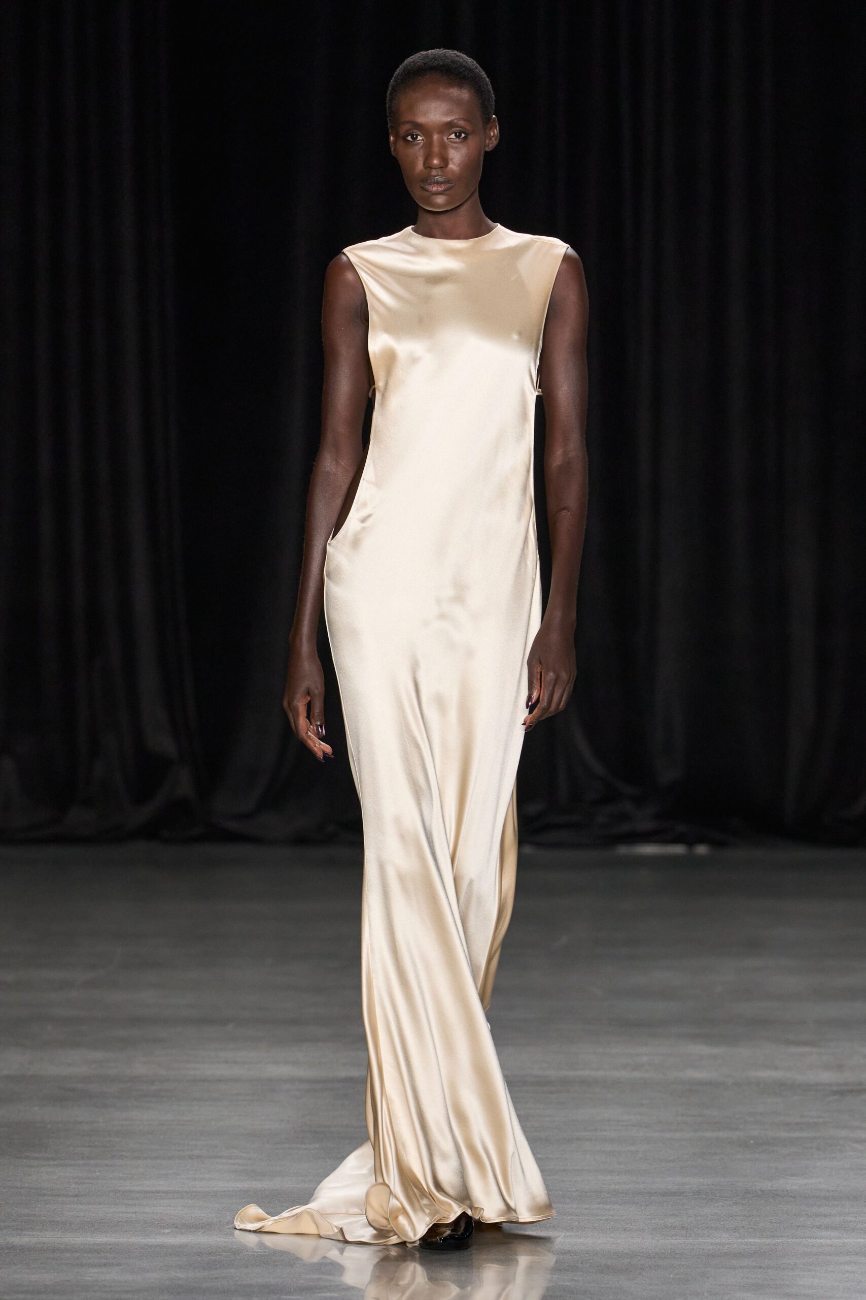 Champagne satin column gown, armholes cut deep and hem pooling into a soft twist. Black patent toes peeking from under the train. For crossing a hotel lobby after midnight with your shoes half hidden.