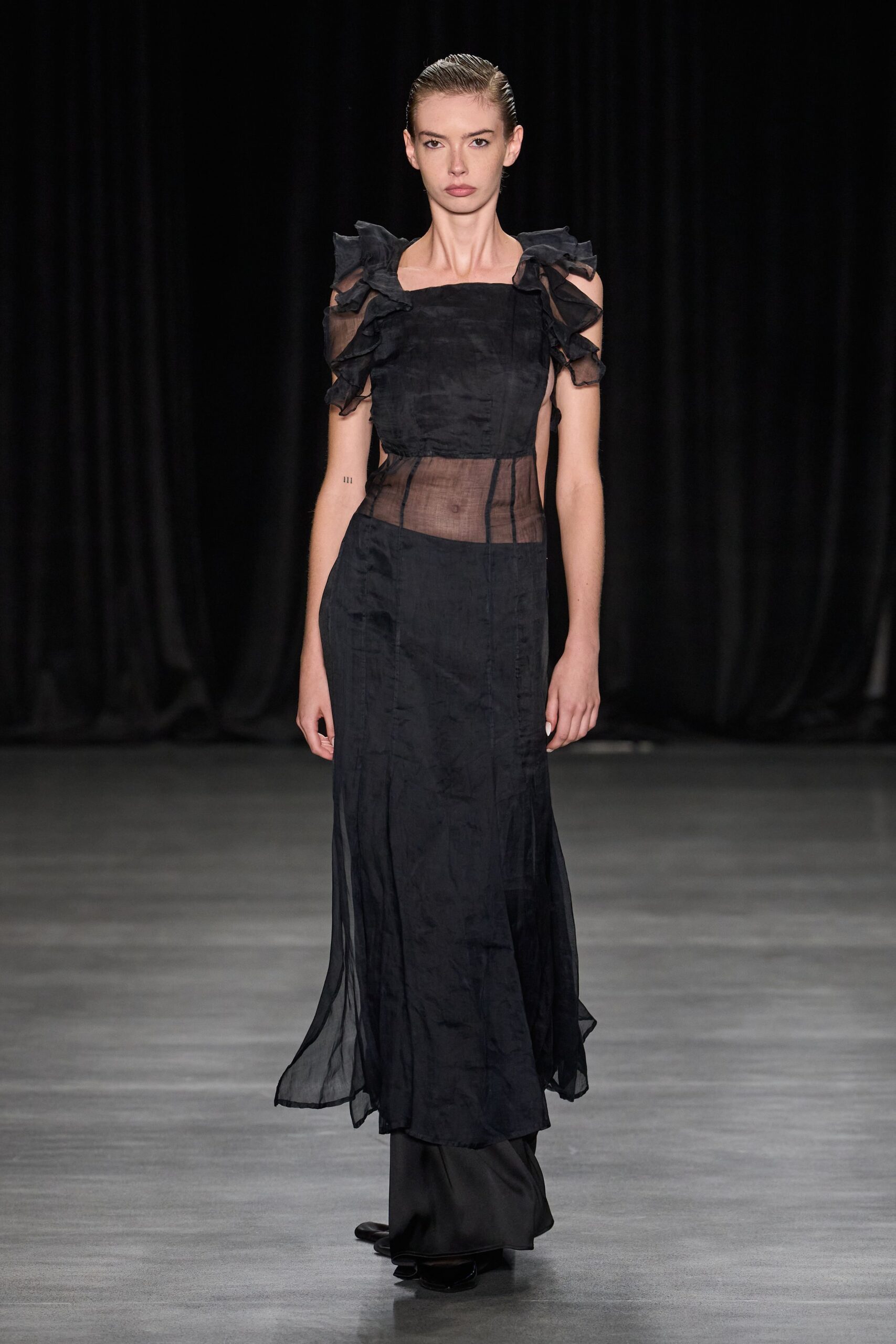 Black organza dress with ruffled shoulder flounces and a sheer waist panel. Black satin trousers pooling under the translucent skirt, hem wavering at the floor. Square-toe black shoes barely breaking the trouser line. Whatever you wear to cross a hotel lobby after midnight for ice.