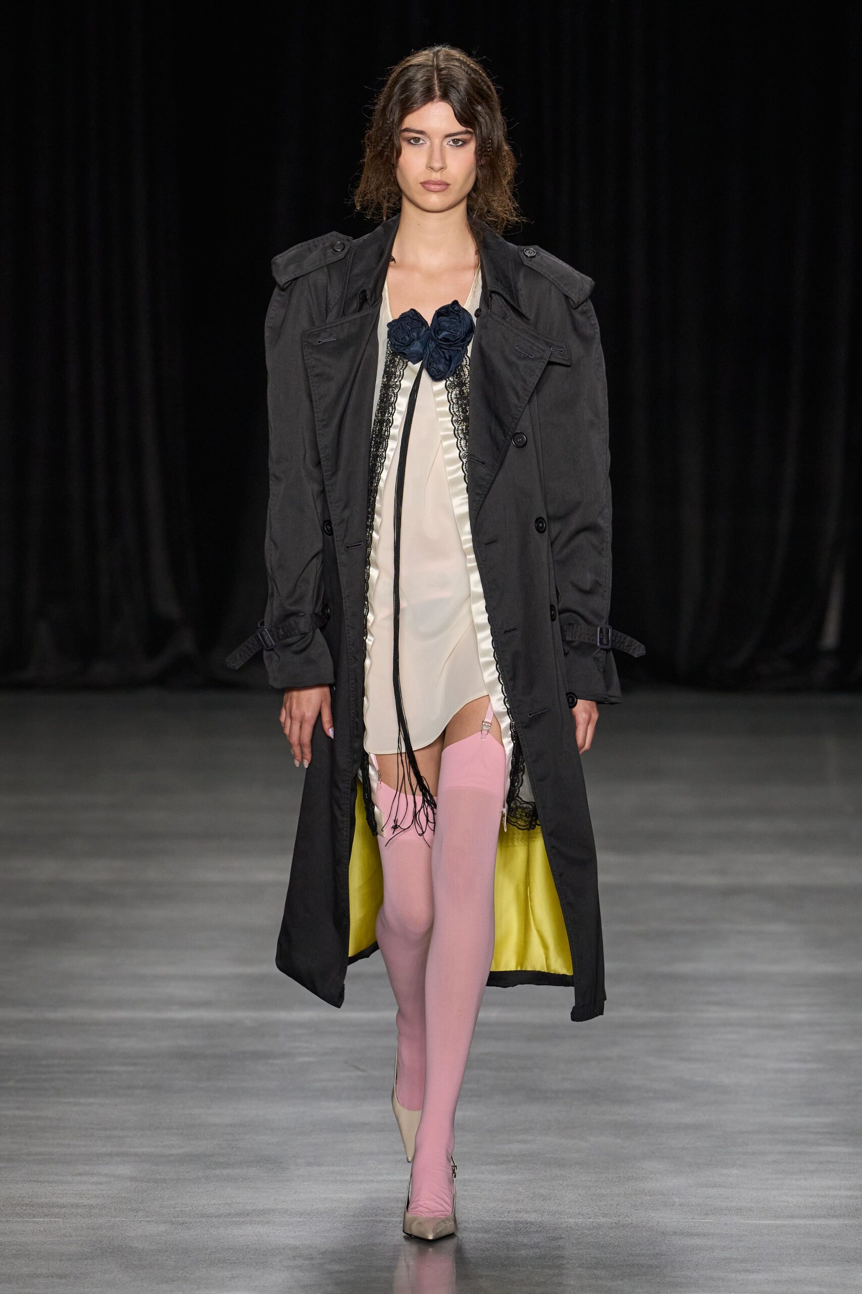 Washed black trench with broad storm flaps and acid-yellow lining. Ivory satin slip edged in black lace, navy rosettes knotted at the chest. Powder-pink thigh-high stockings clipped with visible garter tabs. Nude pointed pumps, one ankle zip glinting. Whatever you wear downstairs when the hotel fire alarm starts at dawn.