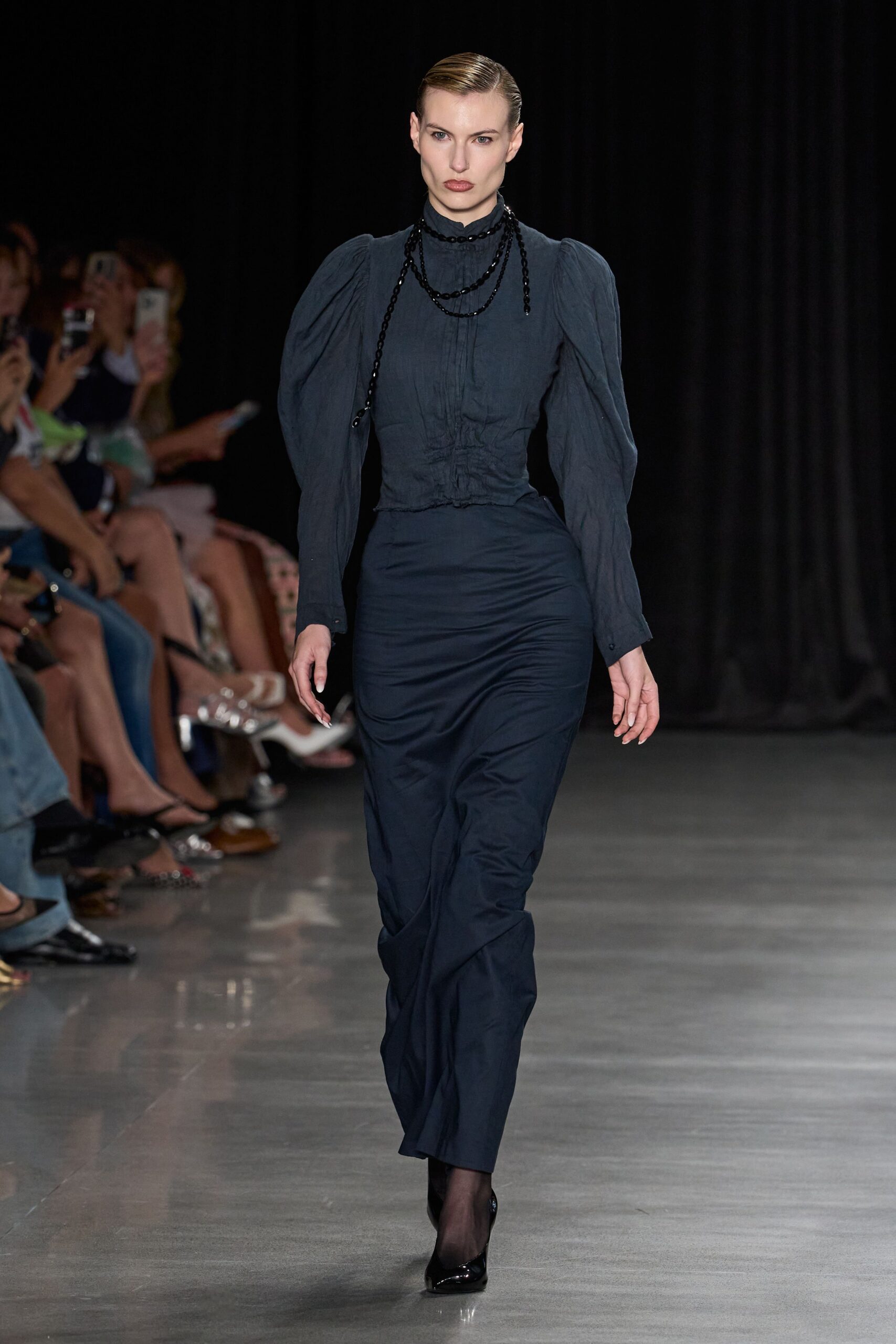 Ink-blue blouse with puffed sleeves and ruched seams pulling tight at the waist. Long black beaded strands looped twice, one end hanging crooked. Dark navy column skirt in smooth matte cloth, lightly wrinkled through the shin. Sheer black tights. Black patent pumps. For reading names from a lectern in a drafty town hall.