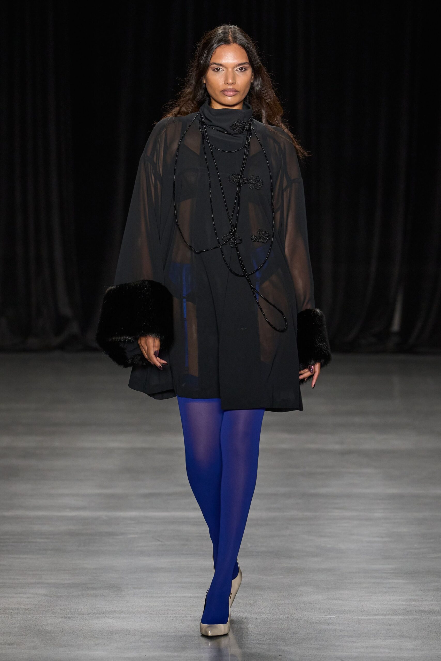 Black sheer chiffon cape with plush fur cuffs swallowing the hands. Black high-neck mini dress with frog closures scattered off-center. Long black beaded cords looping low, one knot hanging near the hem. Opaque cobalt tights. Square-toe oyster leather pumps. Walking from the opera cloakroom into air that still smells of rain.