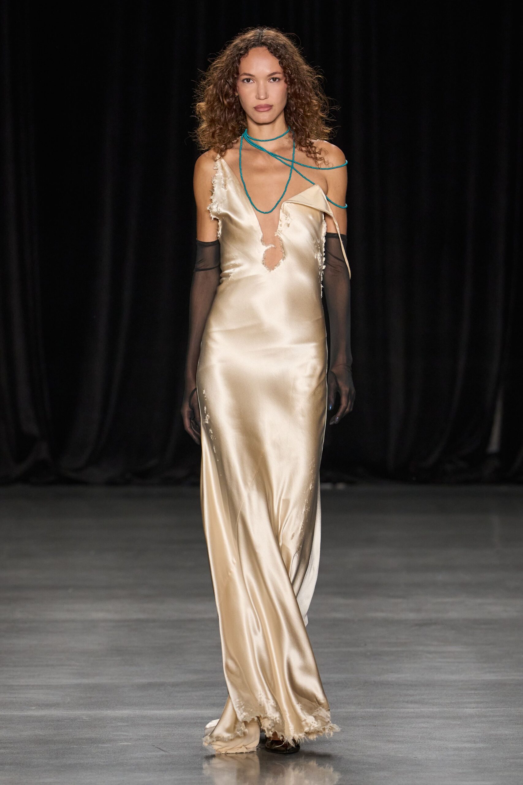 Turquoise bead strands looped twice at the throat and one shoulder. Champagne satin slip dress, plunging keyhole torn with frayed raw edges. Sheer black opera gloves. Dark pointed shoes peeking under the shredded hem. Whatever you wear downstairs when the hotel bar ran past midnight.