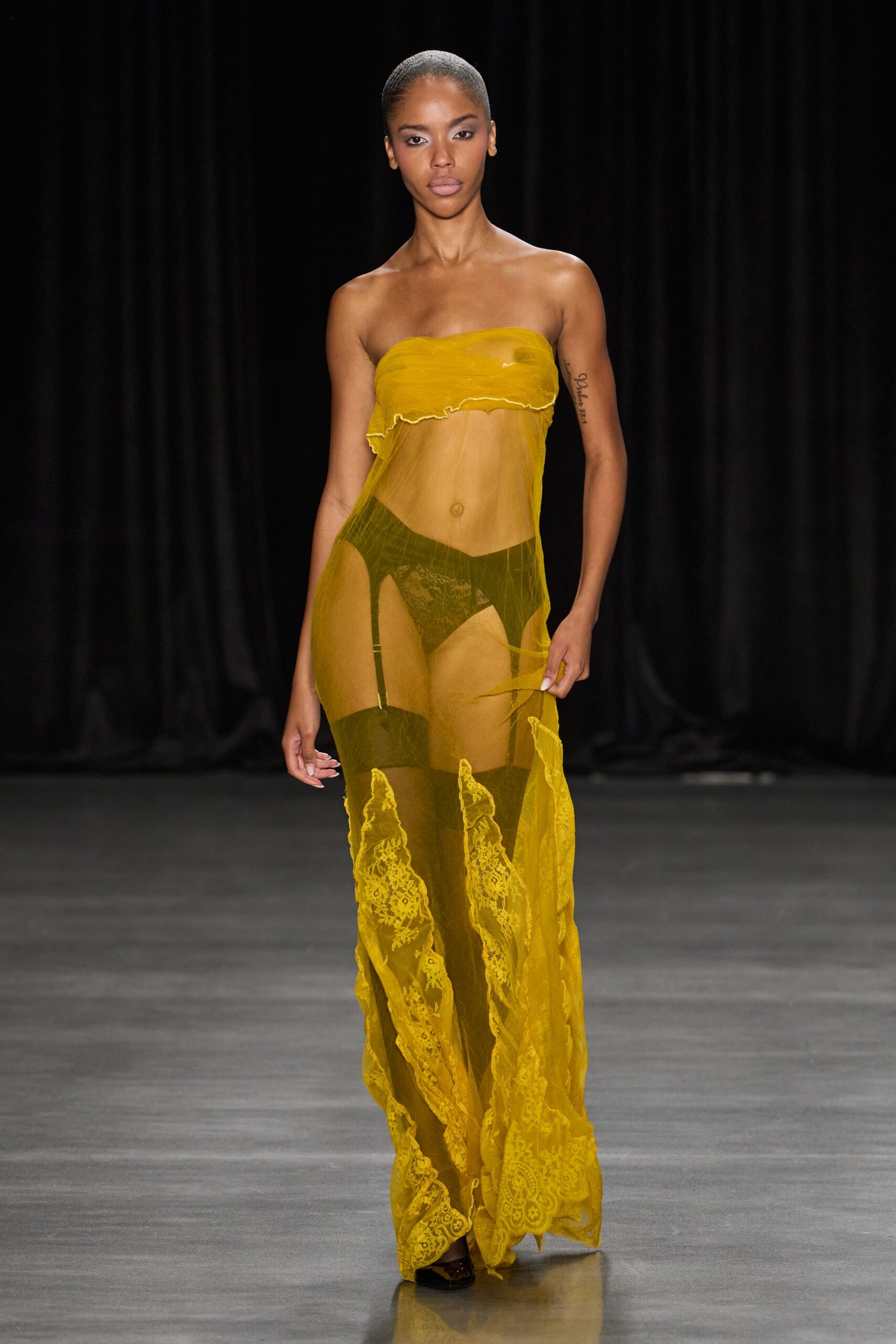 Mustard sheer mesh tube dress, ruched at the bust with raw lettuce edges. Olive lace briefs under a darker V-shaped panel. Black stockings clipped to suspenders, bands stark through the mesh. Dark pointed shoes peeking beneath the lace hem. For crossing a hotel lobby at dawn with last night's makeup still on.