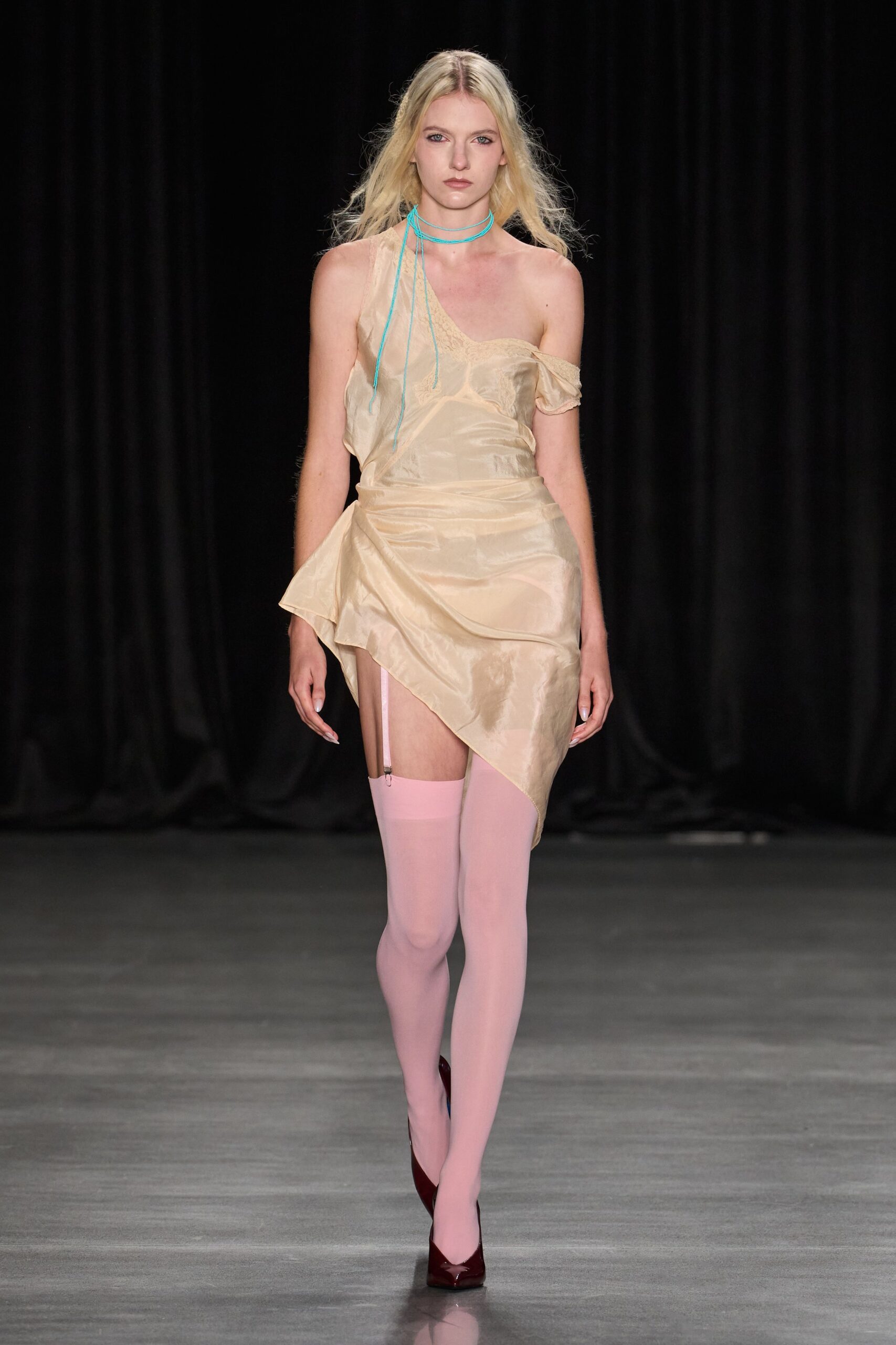 Champagne slip dress in crinkled satin, one strap fallen and hem wrapped askew. Turquoise cord choker trailing loose ends almost to the waist. Powder-pink thigh-highs with a single garter clip left exposed. Burgundy patent pumps with sharp pointed toes. Whatever you pulled back on after dawn, hotel curtains still closed.
