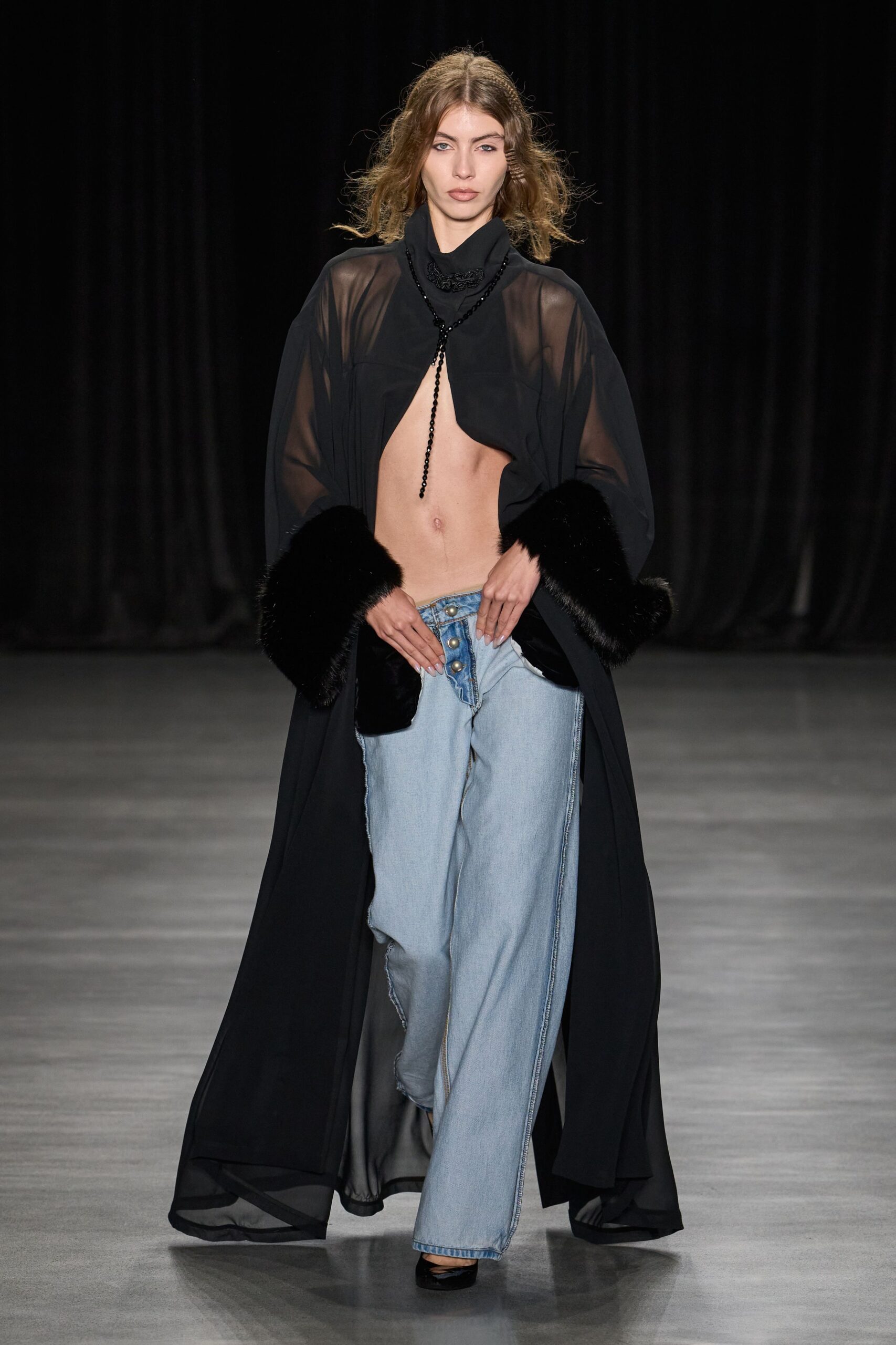 Black sheer robe coat with plush fur cuffs and a dragging hem. High-neck black crop top, cut away to a bare center. Black beaded strand hanging to the navel. Bleached wide-leg jeans, fly left open under three metal buttons. Black patent square toes. Whatever you wear home when dawn finds you under theatre curtains.