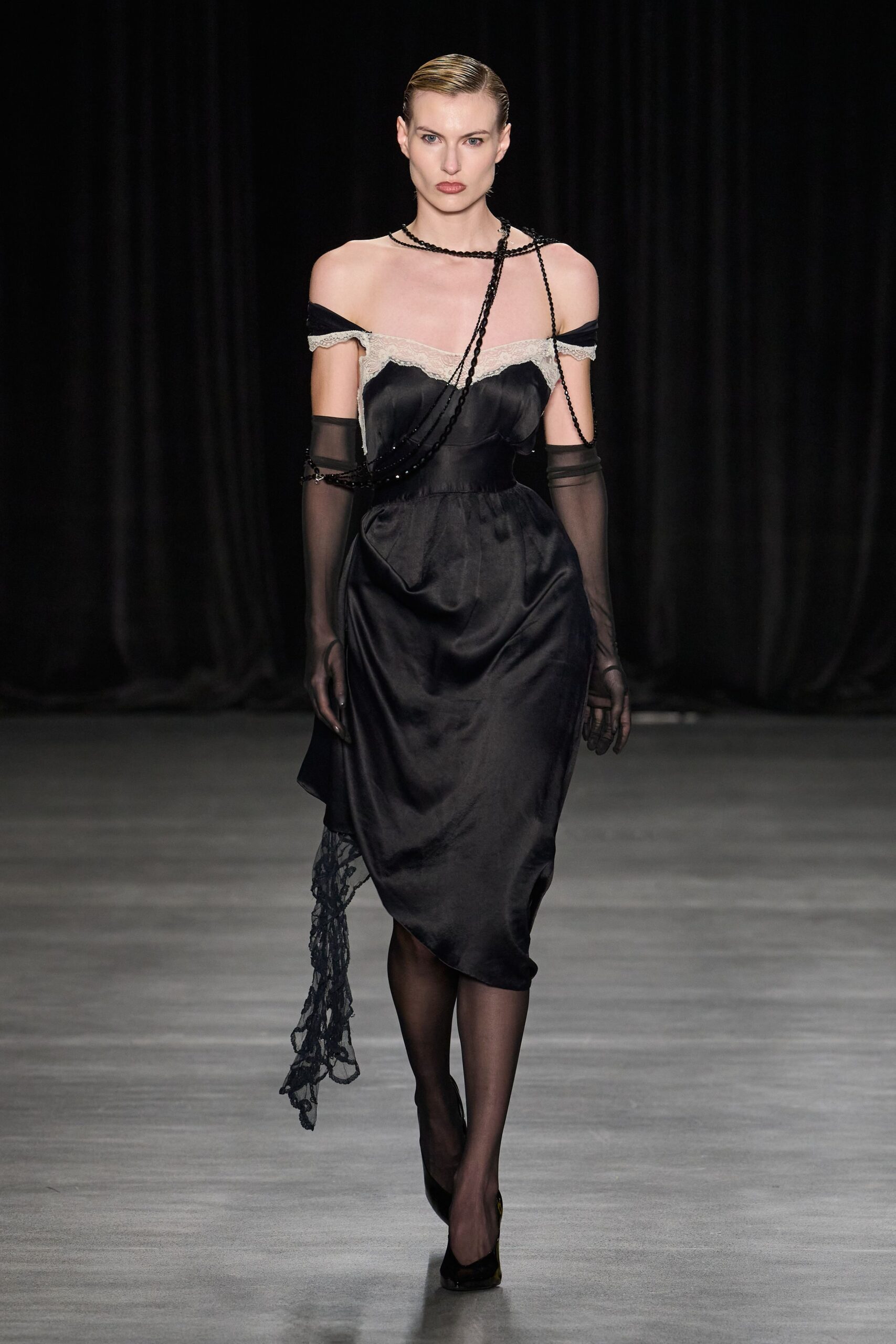 Black satin slip dress with cream lace edging the dropped neckline. Long black bead strands looped across one shoulder and the torso. Elbow-length black mesh gloves, one bracelet snagged at the forearm. Sheer black stockings. Black pumps, glossy at the toe. Whatever you wear downstairs in a grand hotel after missing dinner.