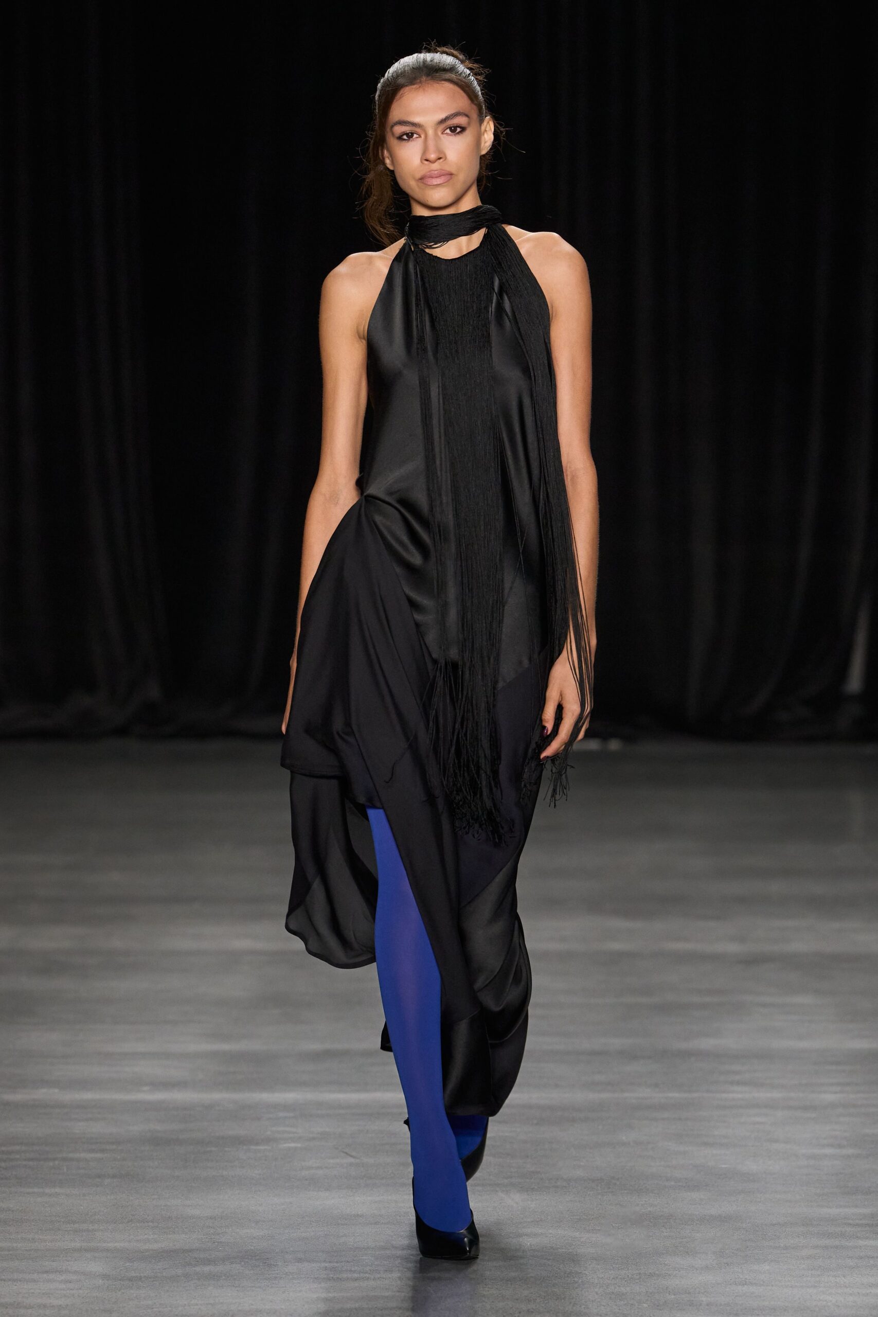 Black crinkled scarf looped at the neck, fringe falling past the waist. Black satin halter dress with a draped handkerchief hem and sheer underlayer. Electric-blue opaque tights slicing through the dark look. Black pointed pumps, glossy at the toe. Crossing a hotel lobby after midnight with one earring missing.