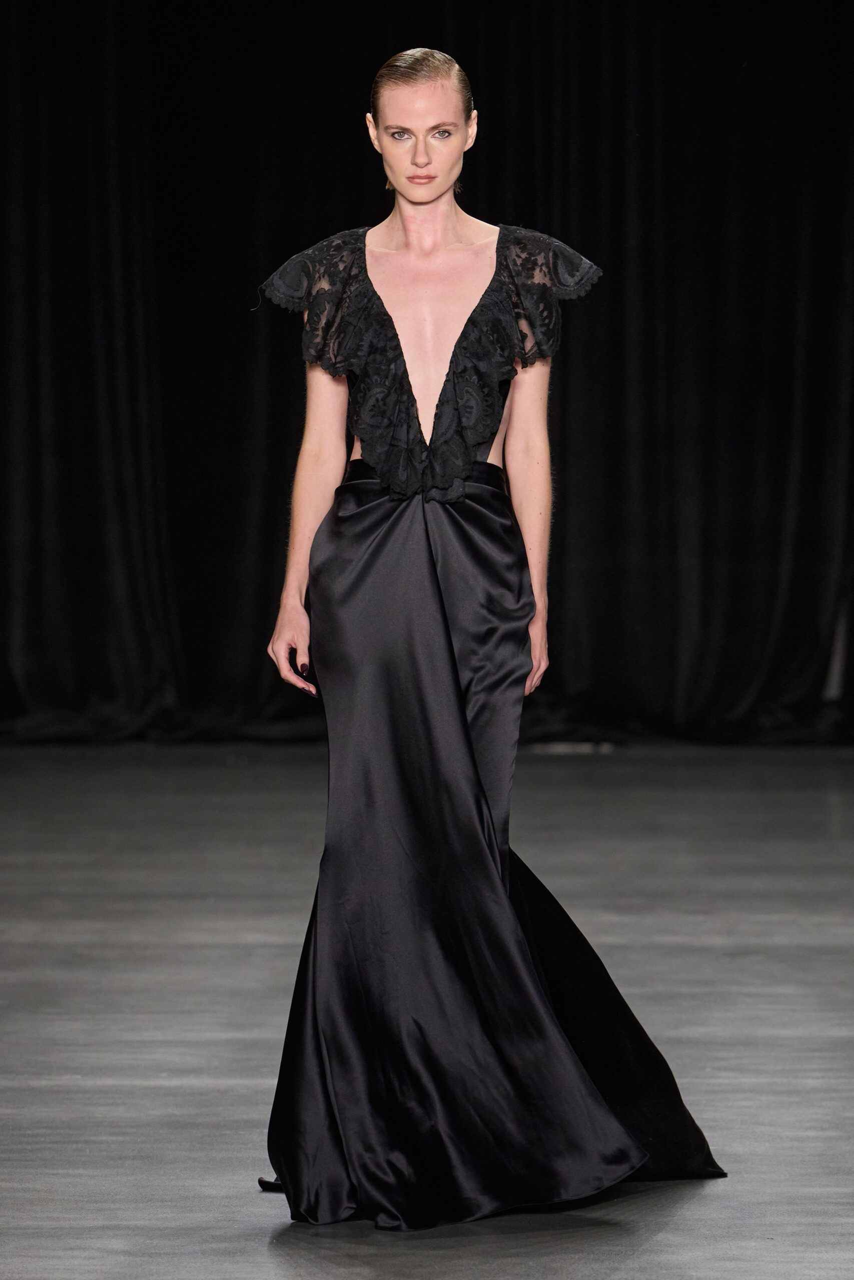 Black satin column gown with a plunging lace bodice and raw flutter sleeves. Crossing a hotel ballroom after midnight, before the chairs are stacked.