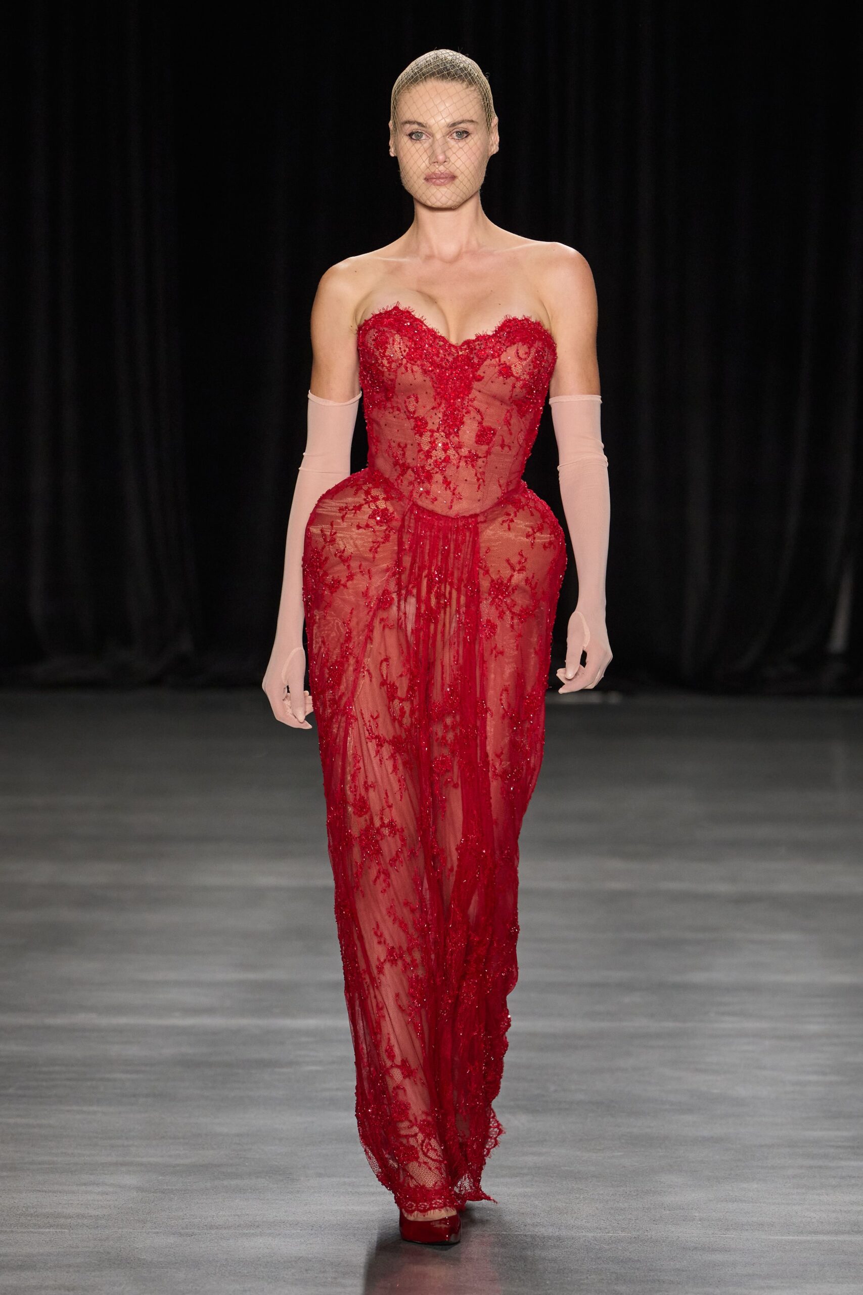 Fine nude netting stretched close over a slicked-back head. Scarlet beaded lace gown, corseted through the sheer nude mesh torso. Pale blush stretch tulle gloves, ending in a small thumbhole. Red satin platforms just visible under the trailing hem. For crossing a hotel ballroom after midnight with the raffle roses still in hand.