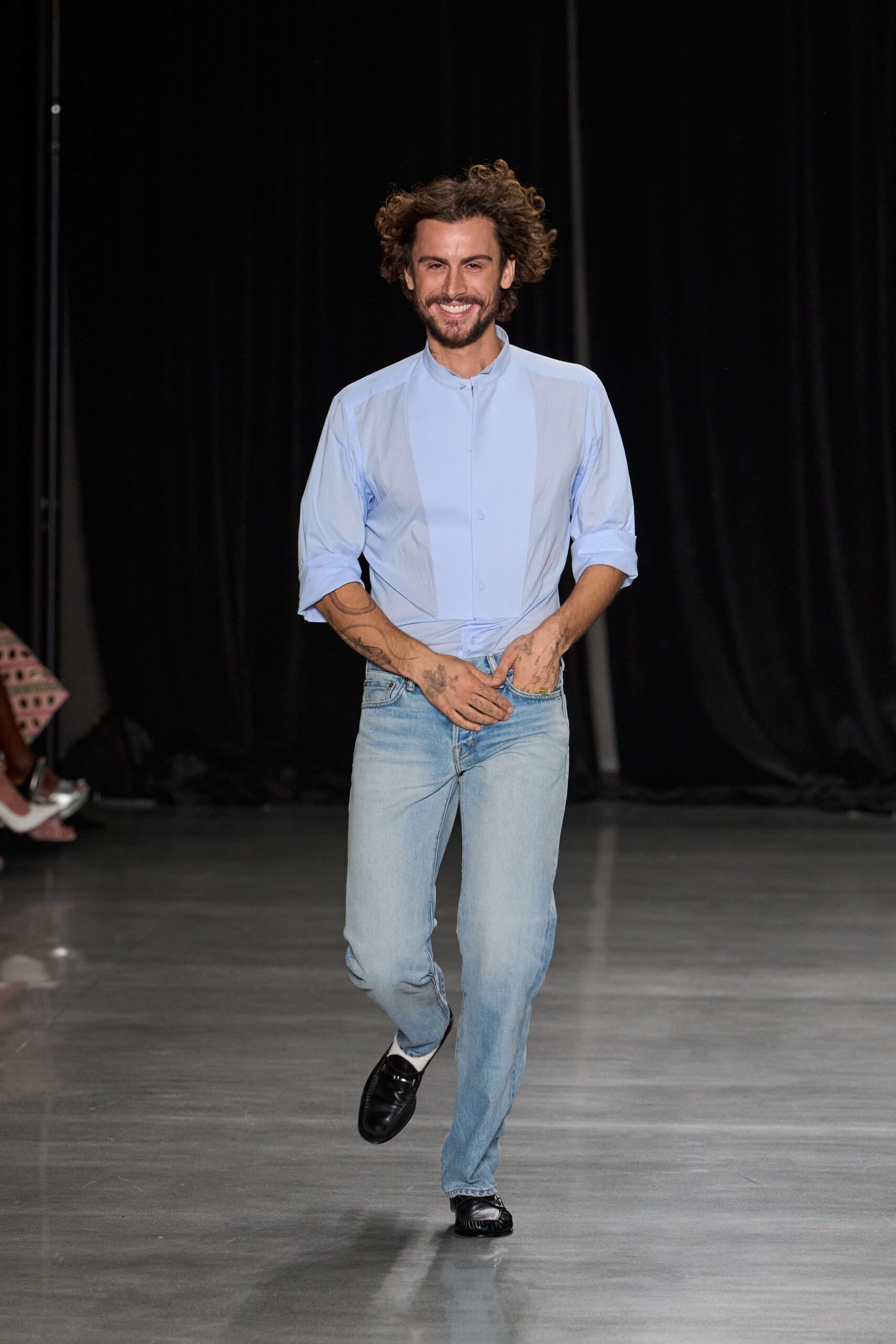 Pale blue poplin shirt with a band collar and hidden placket. Light-wash straight jeans, whiskered at the zip and thigh. White socks flashed above the shoe. Black leather loafers with gathered apron seams. For answering the door on a Sunday with coffee still brewing.