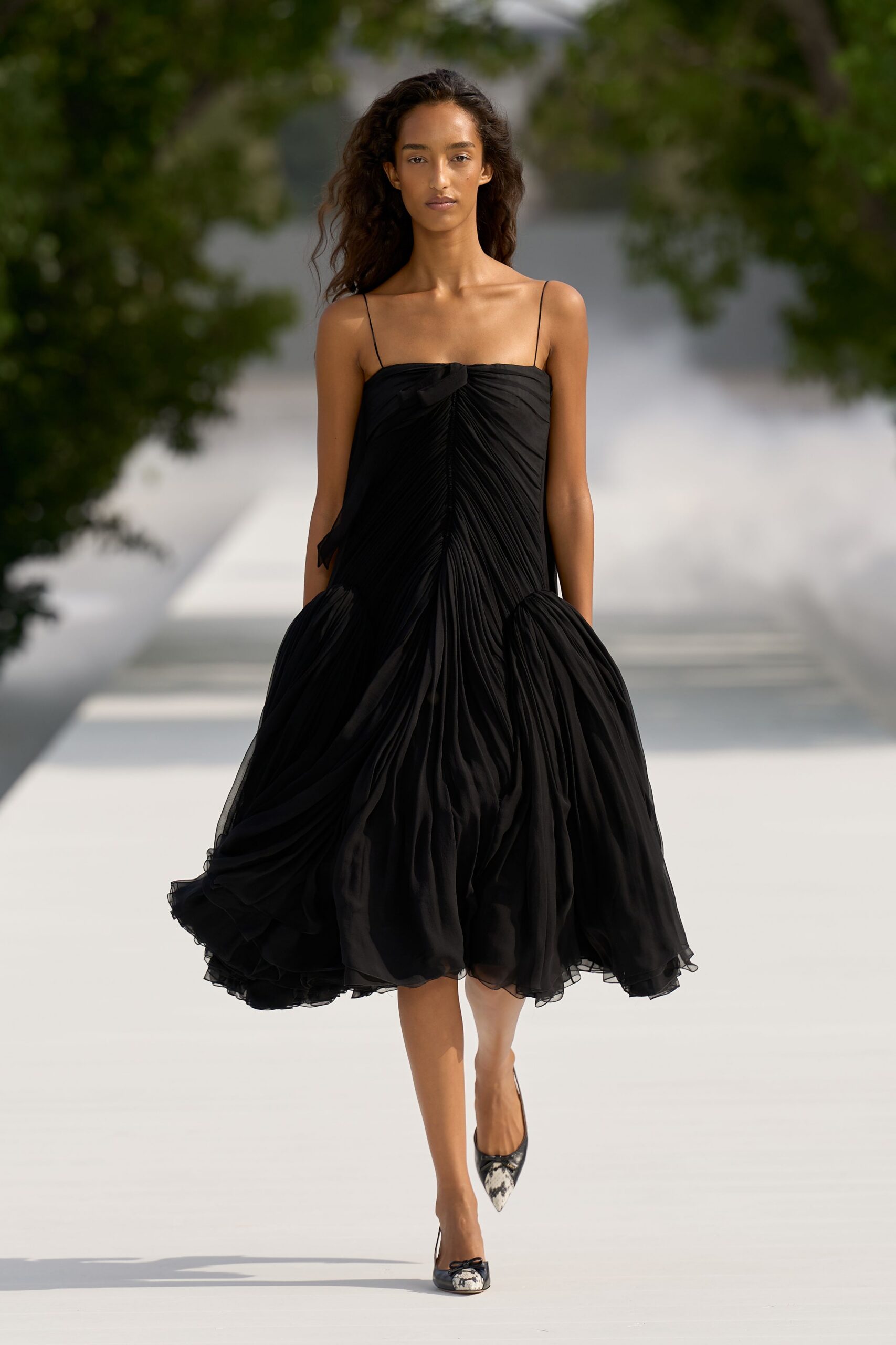 Black chiffon slip dress, knife-pleated through the front into ballooned side panniers. Pointed pumps with mottled snakeskin toes and tiny bow trim. Crossing a white garden path before the wedding guests find their seats.