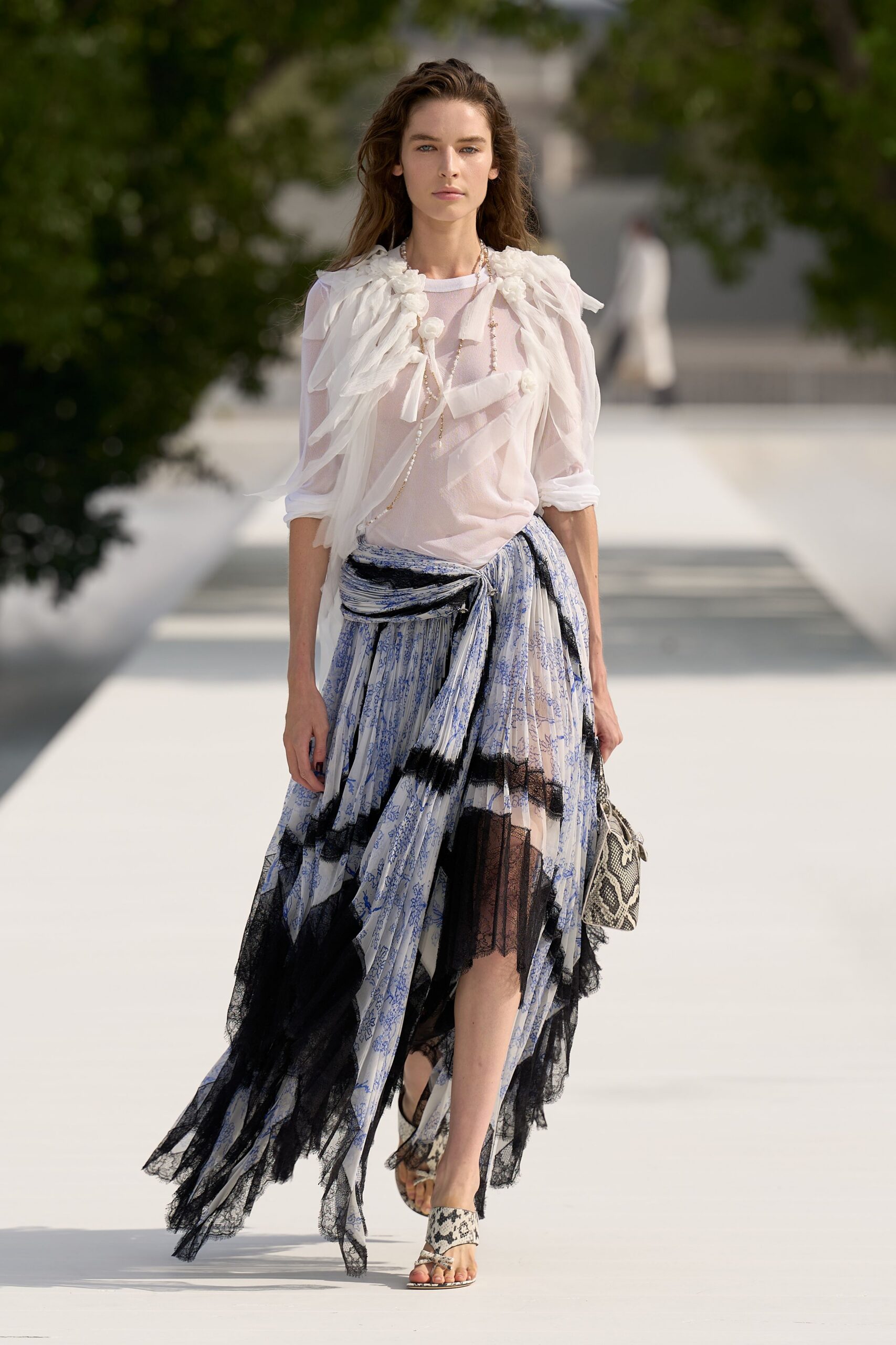 Sheer blush blouse under a froth of petal tulle and rosettes. Long pearl strands with a small tassel slipping off center. Blue toile skirt knotted at the hip, black lace falling ragged. Python bag tucked low. Python thong sandals. Crossing a white courtyard before the garden lunch has fully started.