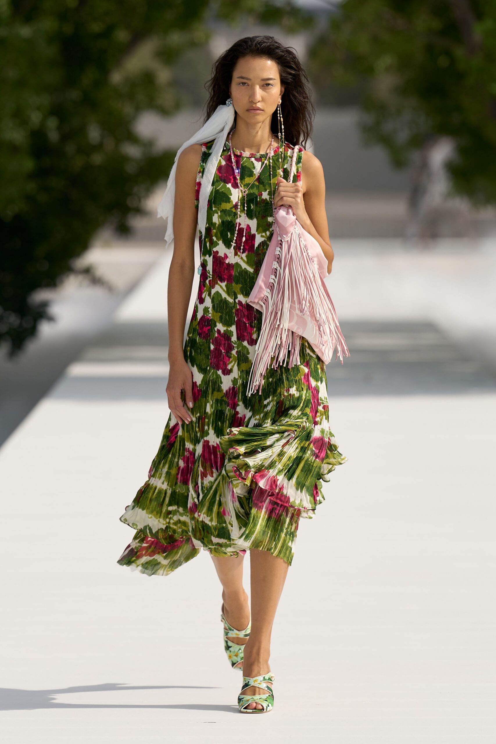 Sleeveless white dress in knife-pleated floral chiffon, hem kicked into ruffles. White gauze scarf knotted at one ear. Blush satin bag with long swaying fringe. Floral mules with crisscross straps. Crossing a hotel courtyard after the wedding brunch ran late.