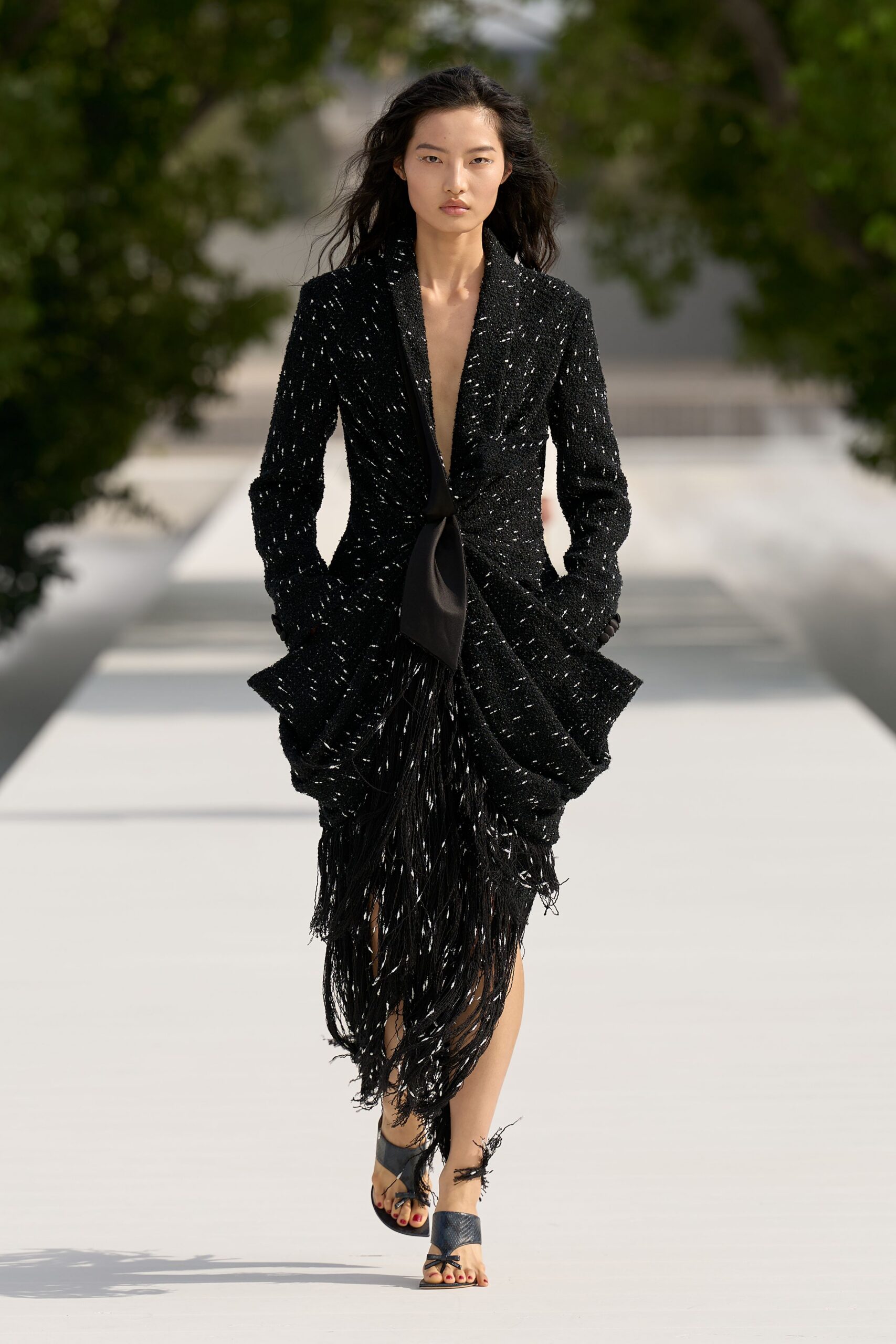 Black sequined tweed dress with plunging shawl lapels and a fringe-caked hem. Satin neck tie knotted low, one blade falling down the center. Black thong sandals, frayed ankle ties whipping loose at the shin. Crossing a sunstruck courtyard after the wedding band has packed up.