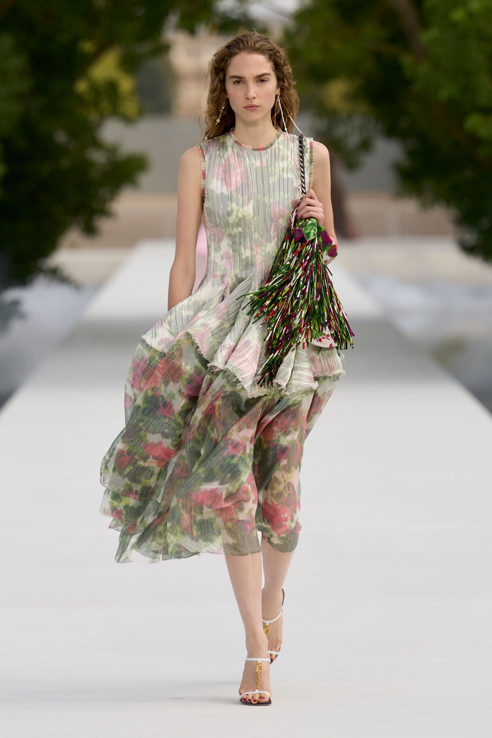 Watercolor floral chiffon dress, finely plissed with a frayed floating peplum. Long pearl-drop strands brushing the shoulders. Multicolored ribbon-fringe bag swinging from a braided strap. White ankle-strap sandals with a dangling gold initial charm. Walking through a palace garden after the sprinkler mist has settled.