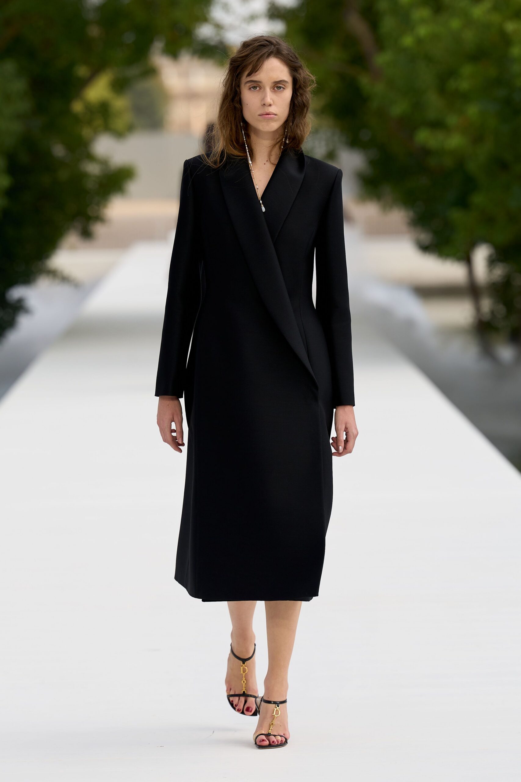 Black wool coatdress cut with sharp shoulders and a long shawl lapel. Fine beaded lariat dropping past the V with a tiny pearl tip. Black ankle-strap sandals with dangling gold letter charms at the vamp. For crossing a palace courtyard after dark, fountain water carrying the sound.