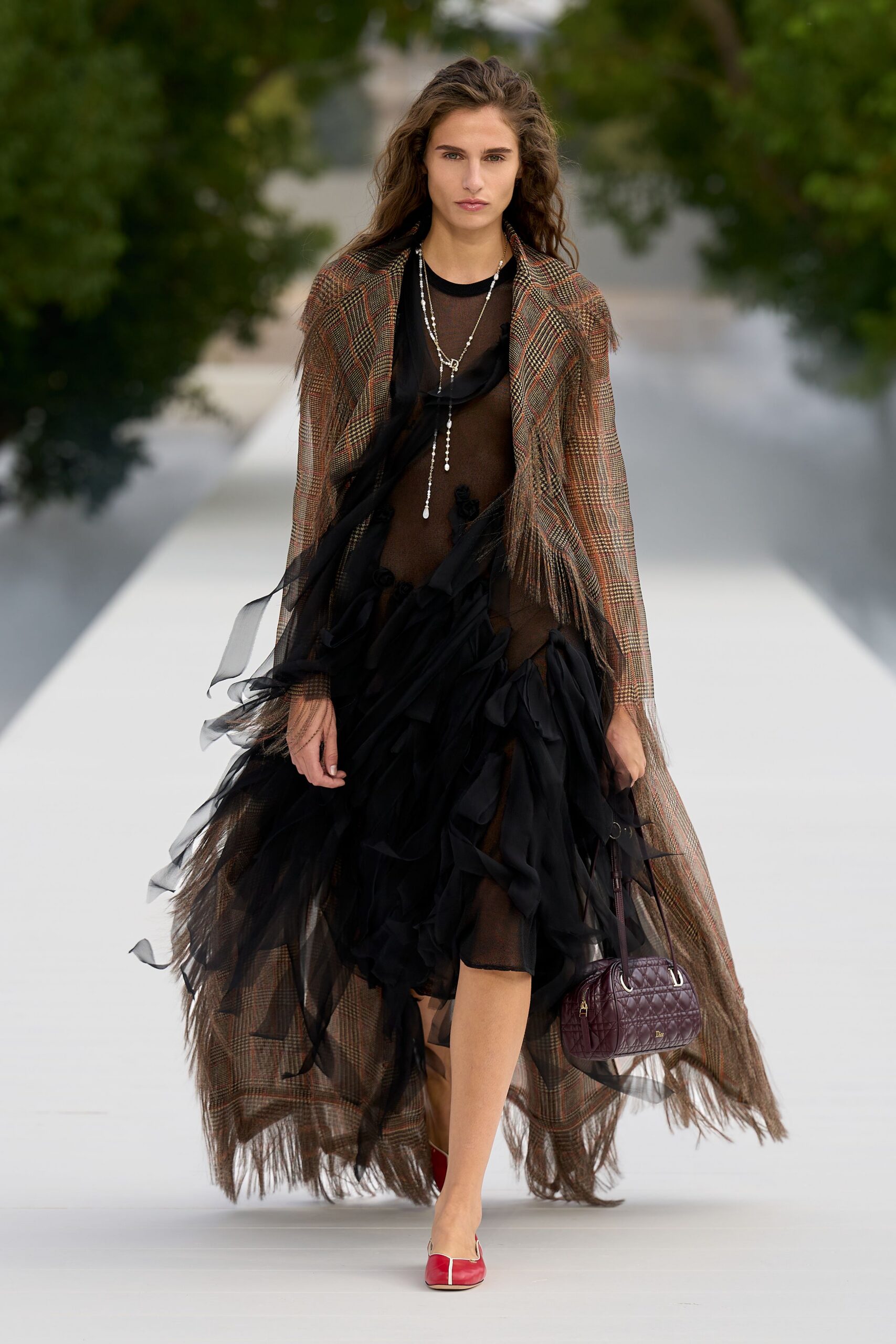 Brown glen-plaid cape coat, long fringe brushing the calves. Sheer black mesh dress swarmed with loose chiffon ruffles. Long silver drop necklace. Burgundy quilted barrel bag with ring handles. Red flats split by a pale center seam. Crossing a hotel garden path before the wedding guests find you.