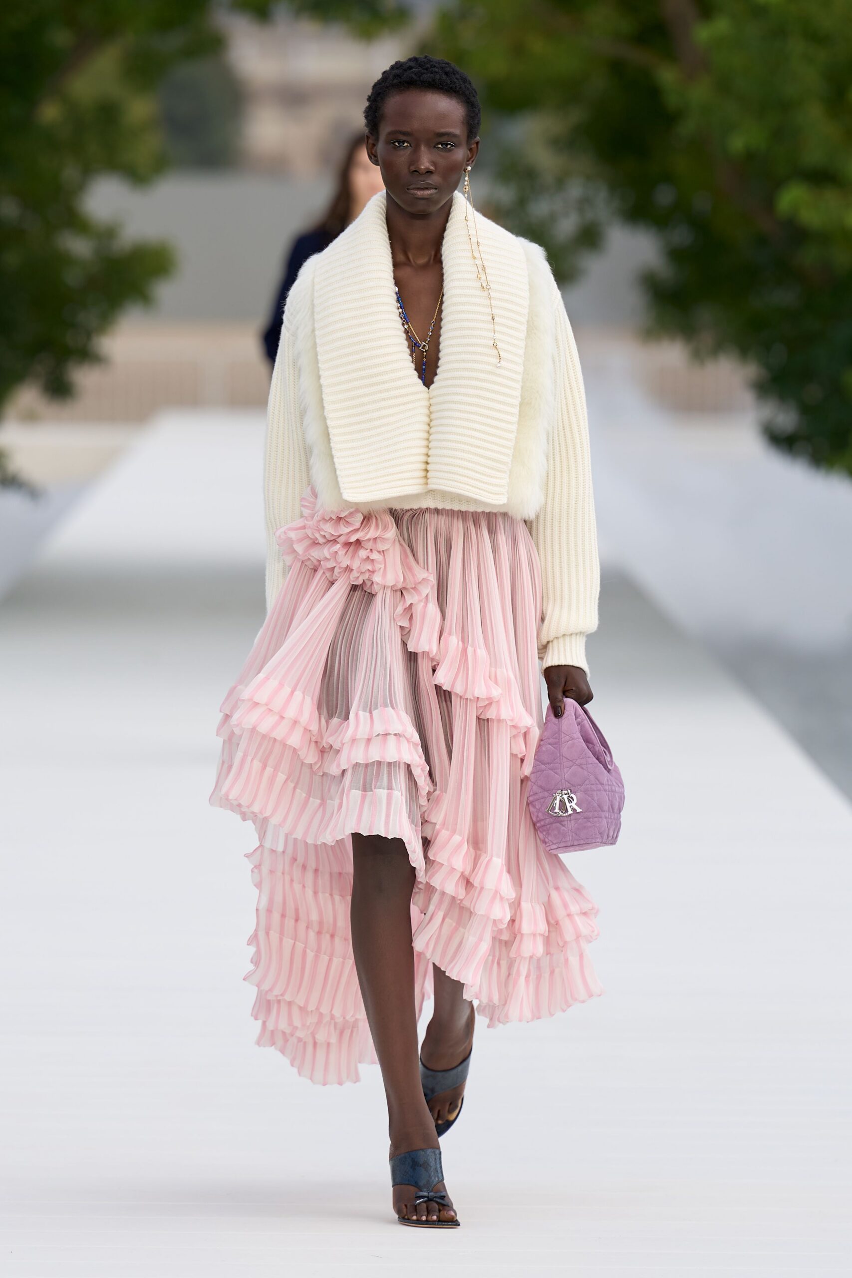 Cream ribbed cardigan with a deep shawl collar and furry body. Blush chiffon skirt, knife-pleated and tiered with striped ruffle flounces. Lilac quilted pouch bag with silver initials. Slate satin mules, tiny bow at the toe. Walking the garden aisle after the cake has already been cut.
