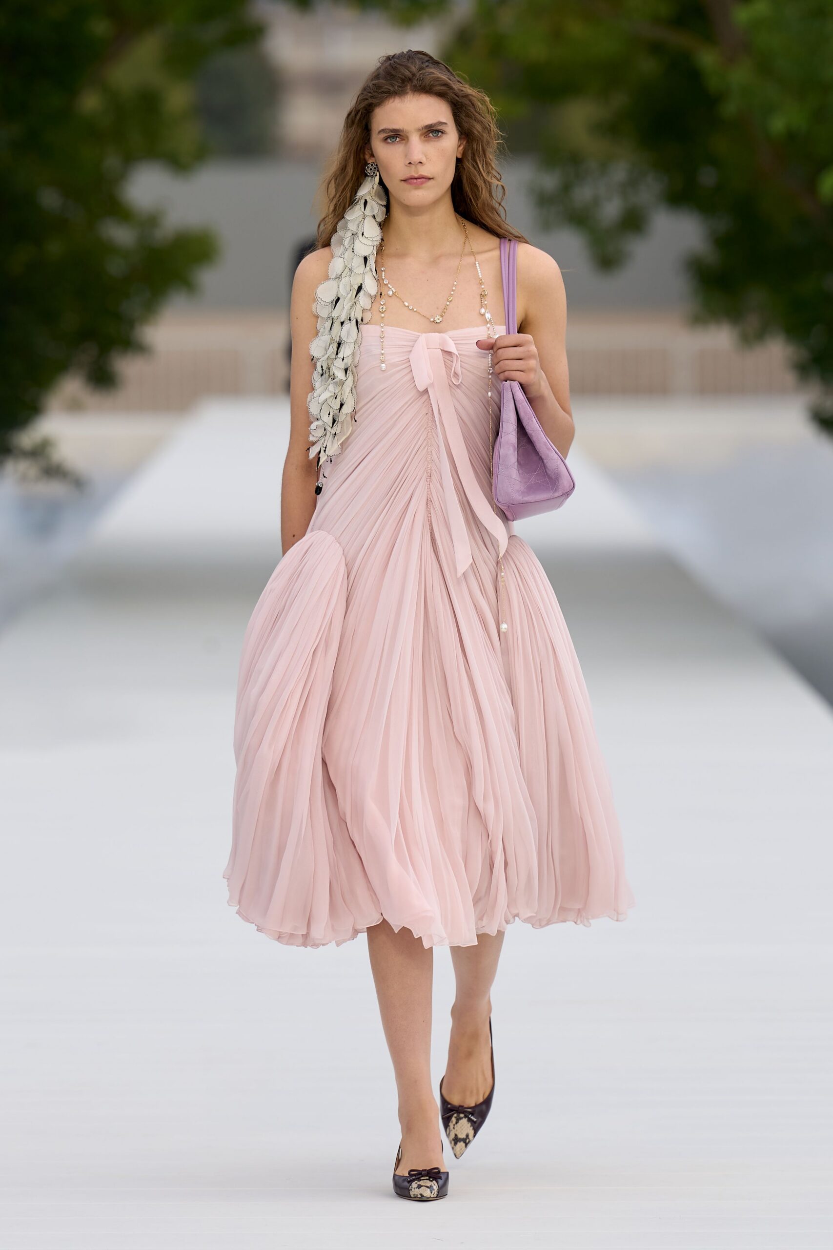 Long cascade of white petal discs, dark whipstitch edging each one. Blush chiffon dress, finely pleated with a tied bow at the bust. Pearl strands dropping low. Lilac shoulder bag in cracked leather. Brown pumps with python toe caps and tiny bows. Garden wedding, halfway through the champagne before dinner.