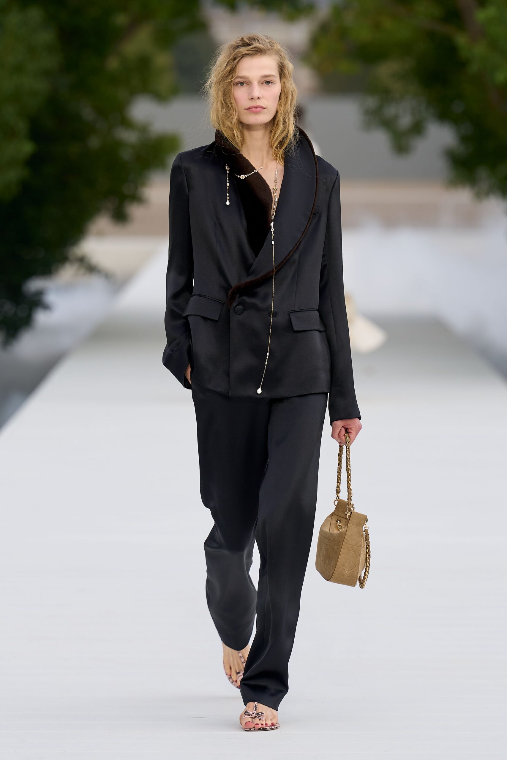 Black satin blazer, one lapel edged in dark mink and cut plunging. Long pearl-drop chains trailing past the navel. Fluid black trousers breaking softly over the foot. Beige quilted shoulder bag with a chunky chain strap. Python-print thong sandals. Garden reception just before sunset, drinks sweating onto the tray.