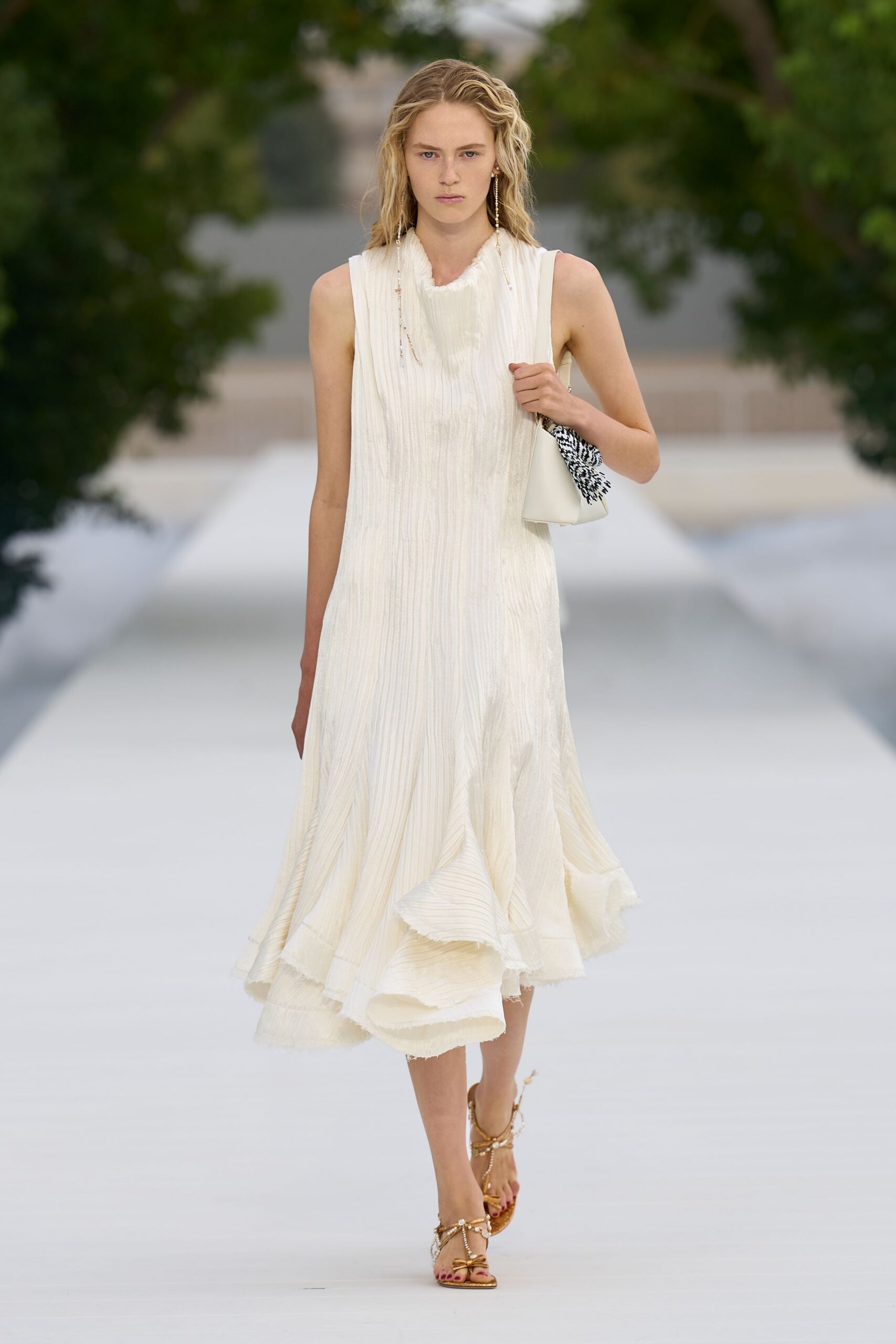 Ivory crinkled dress with a softly ruched funnel neck and raw flounced hem. Long beaded strands brushing the shoulders. Cream shoulder bag with a black-and-white tasselled scarf tied on. Gold thong sandals laced in pearl-strung cords. Walking from the ceremony to the lawn before anyone finds the cake.