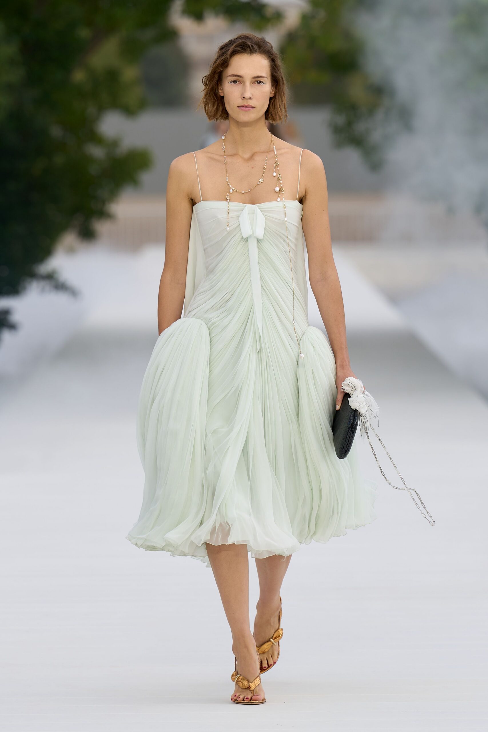 Pale celadon chiffon dress, sunburst pleats gathered to a soft bow at the bust. Long pearl chain looping low past the waist. Black oval clutch with a white scarf knotted round the handle. Gold thong sandals with rounded knot hardware. Crossing a limestone courtyard just before the wedding dinner starts.