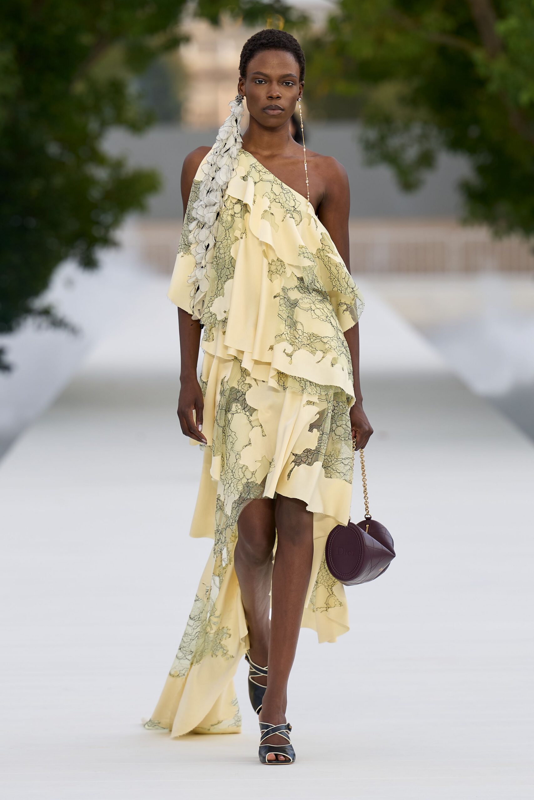 One beaded shoulder-grazer, one small silver stud. Buttercream one-shoulder chiffon dress, black lace print and tumbling ruffles. Plum round bag on a gold chain. Black heeled sandals with thin ivory crisscross straps. Garden ceremony drifting into dusk before anyone calls for cars.